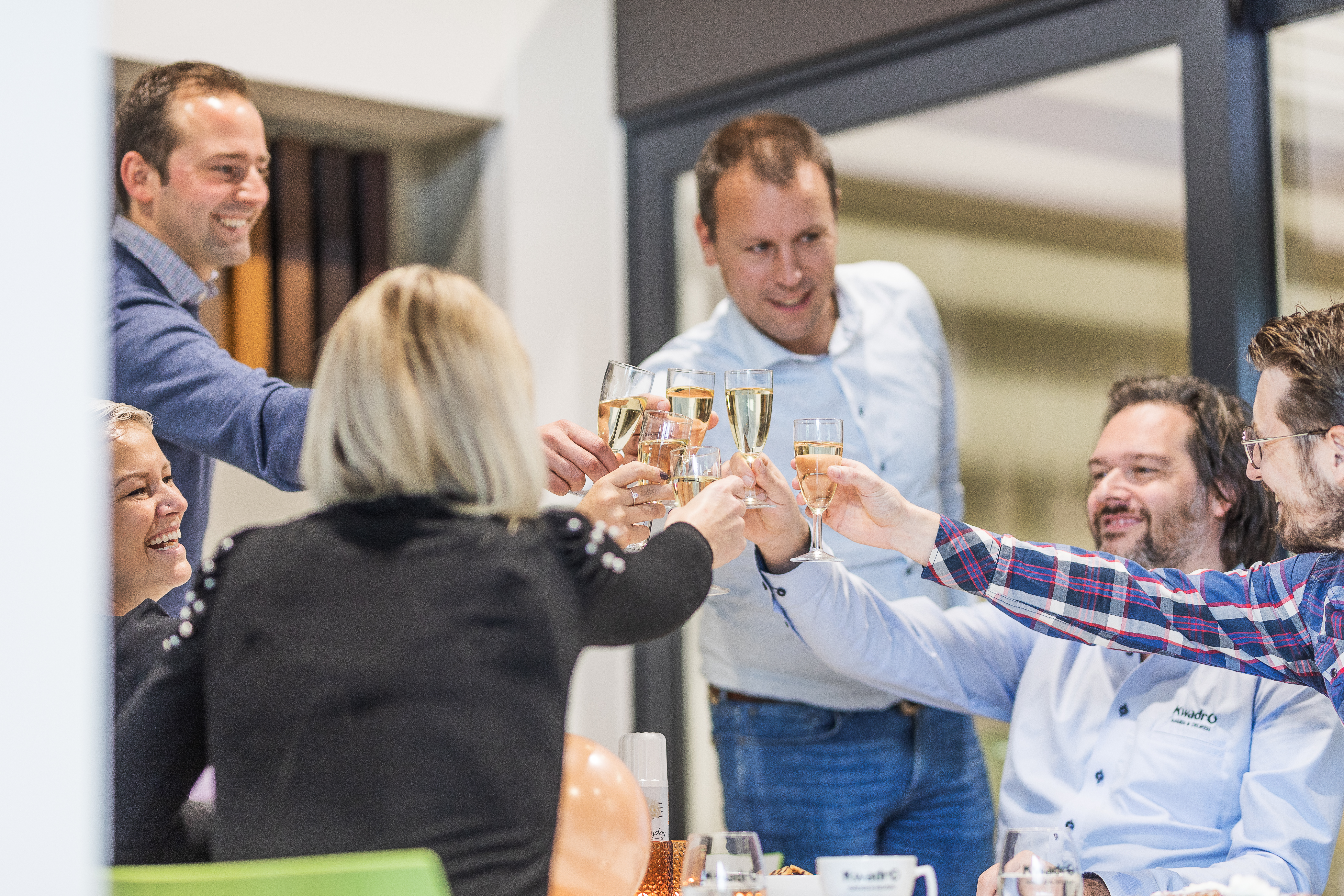 Meerdere personen klinken met glazen aan in een lichte binnenruimte, voor een grote raampartij met donkere raamprofielen.