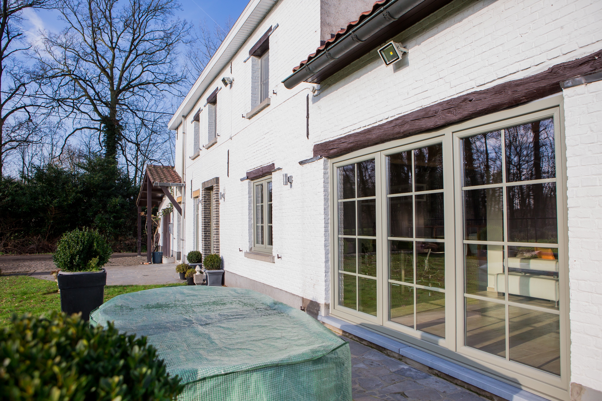 Betongrijs raam met driedubbele beglazing en grote glaspartijen in de gerenoveerde gevel van een vrijstaande woning met een gemoedelijke, tijdloze uitstraling.
