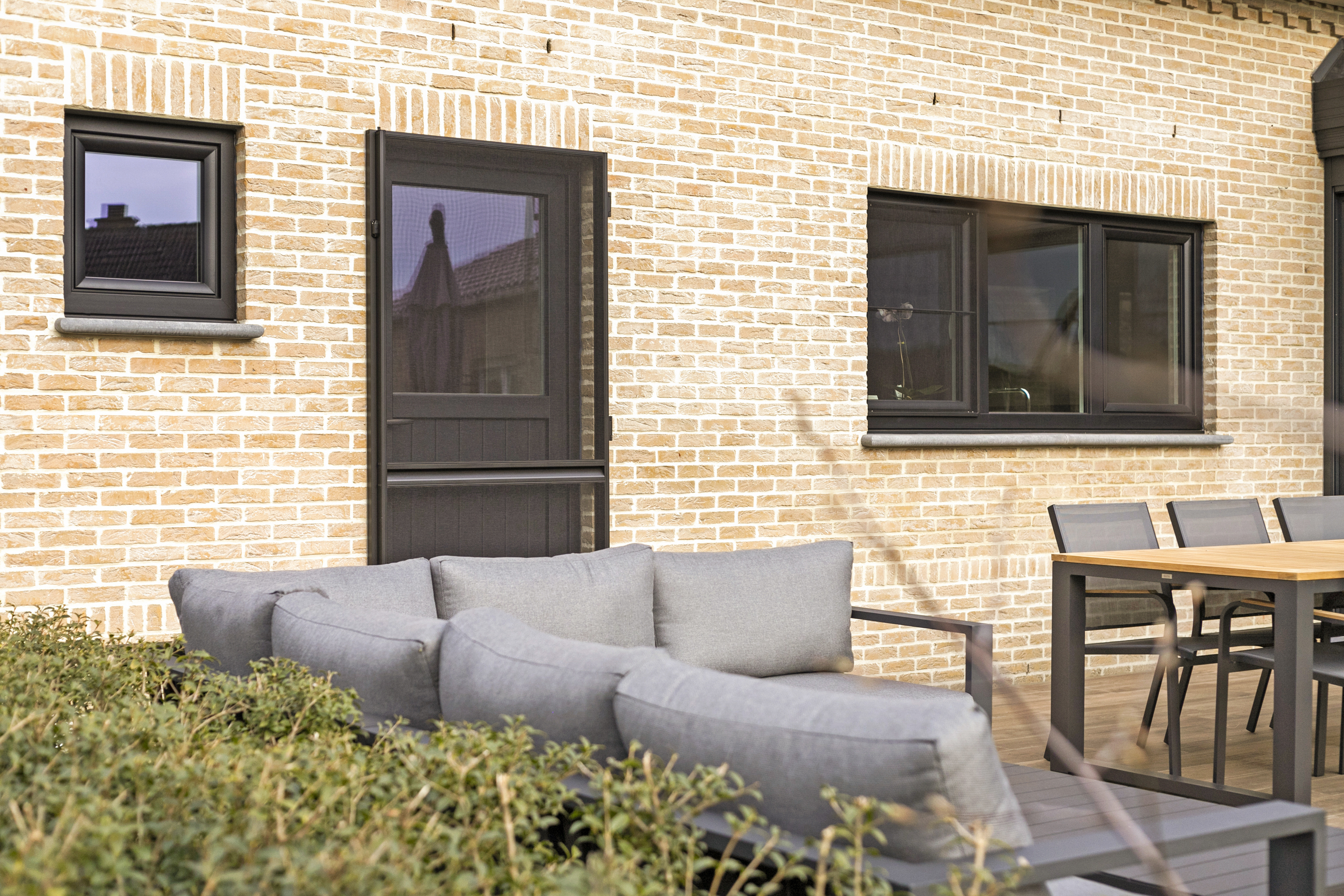 Bakstenen gevel met zwarte matte PVC Impakt PLUS-ramen, een raam met drie verticale delen en draai-kipraam, en een paneeldeur met semi-beglazing en vliegendeur naast terrasmeubilair.