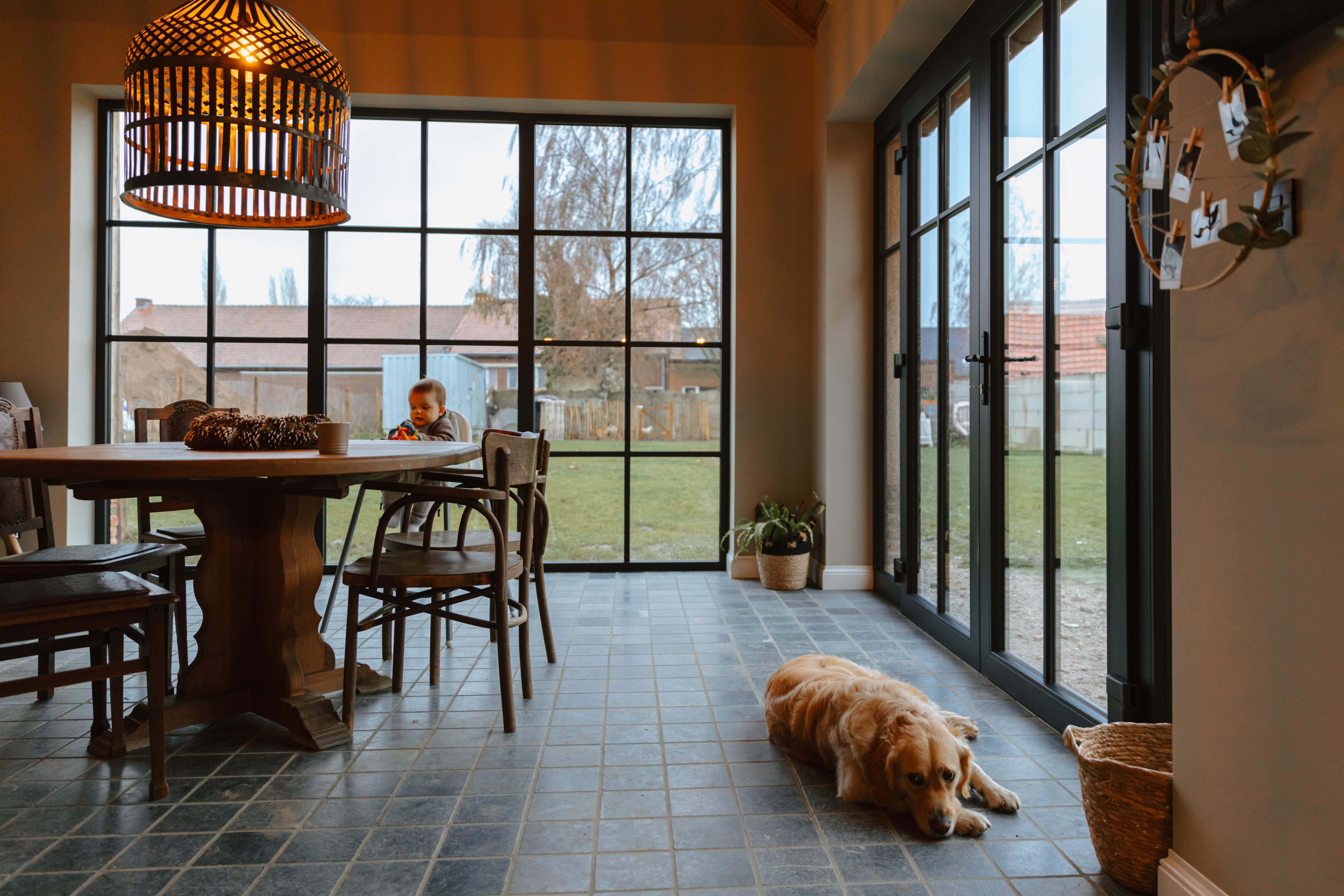 Een persoon zit aan de eettafel in een warme, landelijke woning met driedubbele beglazing en een dubbele glazen deur; een hond ligt op de tegelvloer.