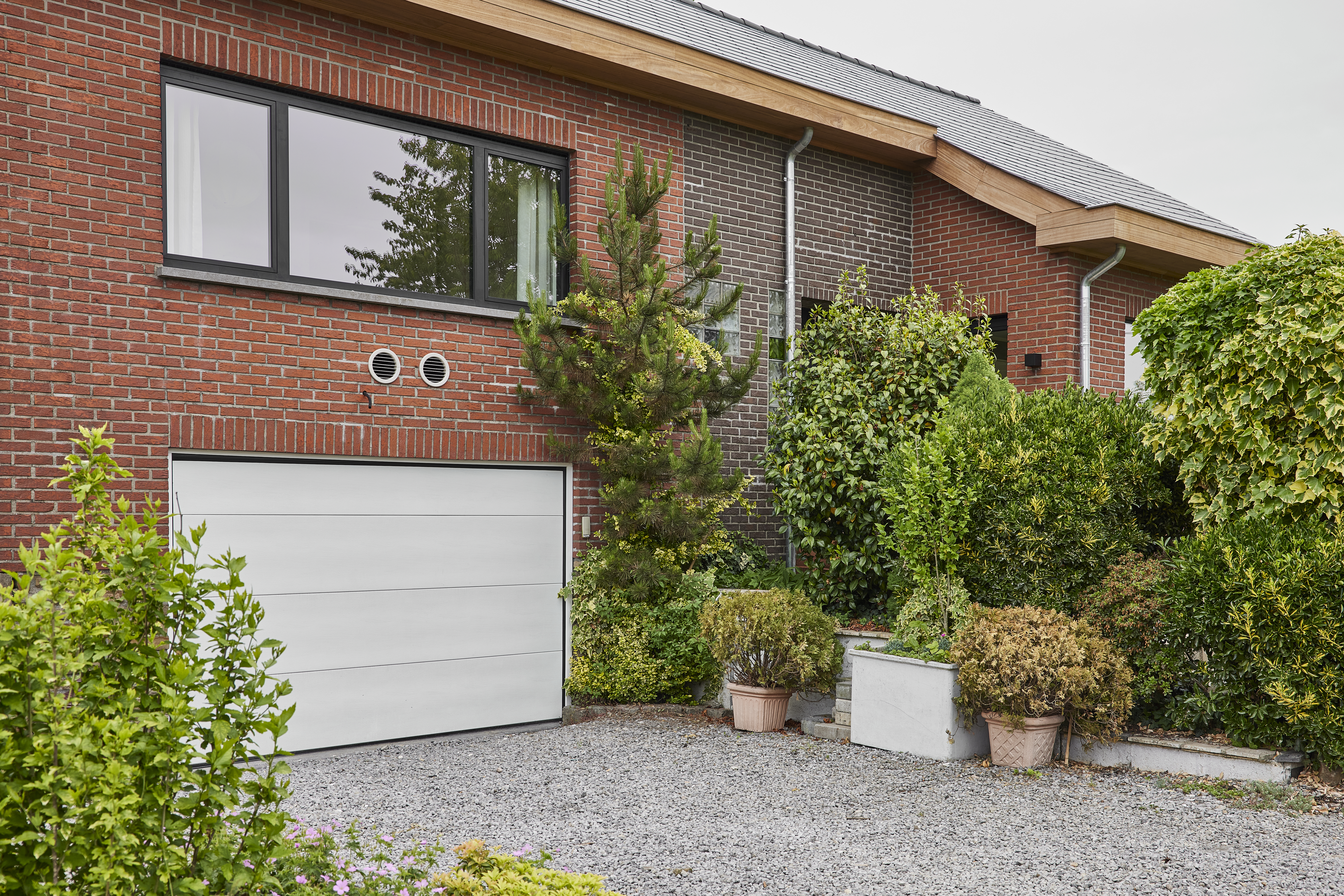 Grafietzwart Komfort EVO small-raam met een vast deel, een draai-kipdeel en drie verticale delen in een bakstenen gevel boven een garage.