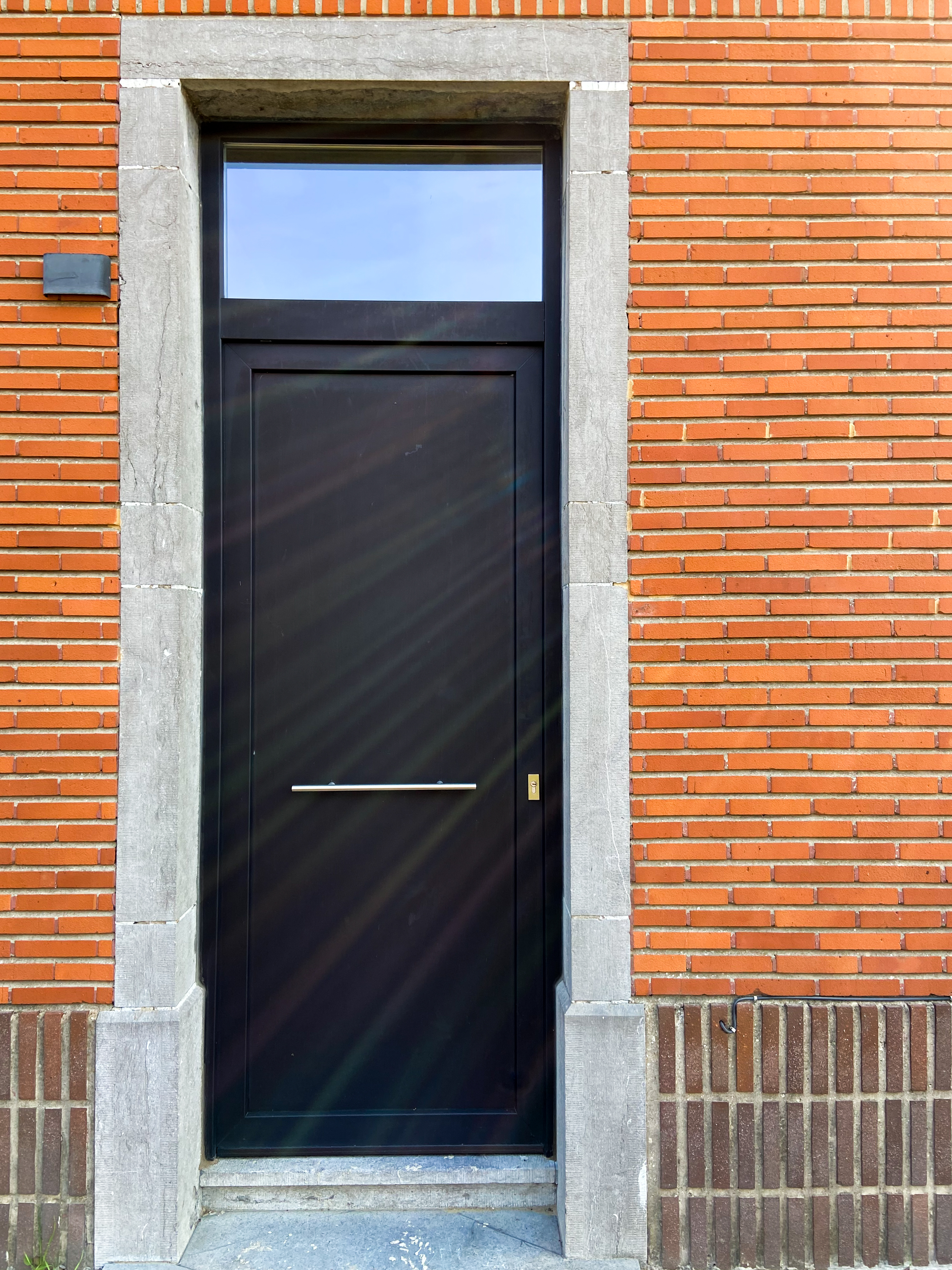 Zwarte aluminium voordeur met bovenlicht en paneel, omlijst door natuursteen in de rode bakstenen gevel van een gerenoveerde vrijstaande woning.