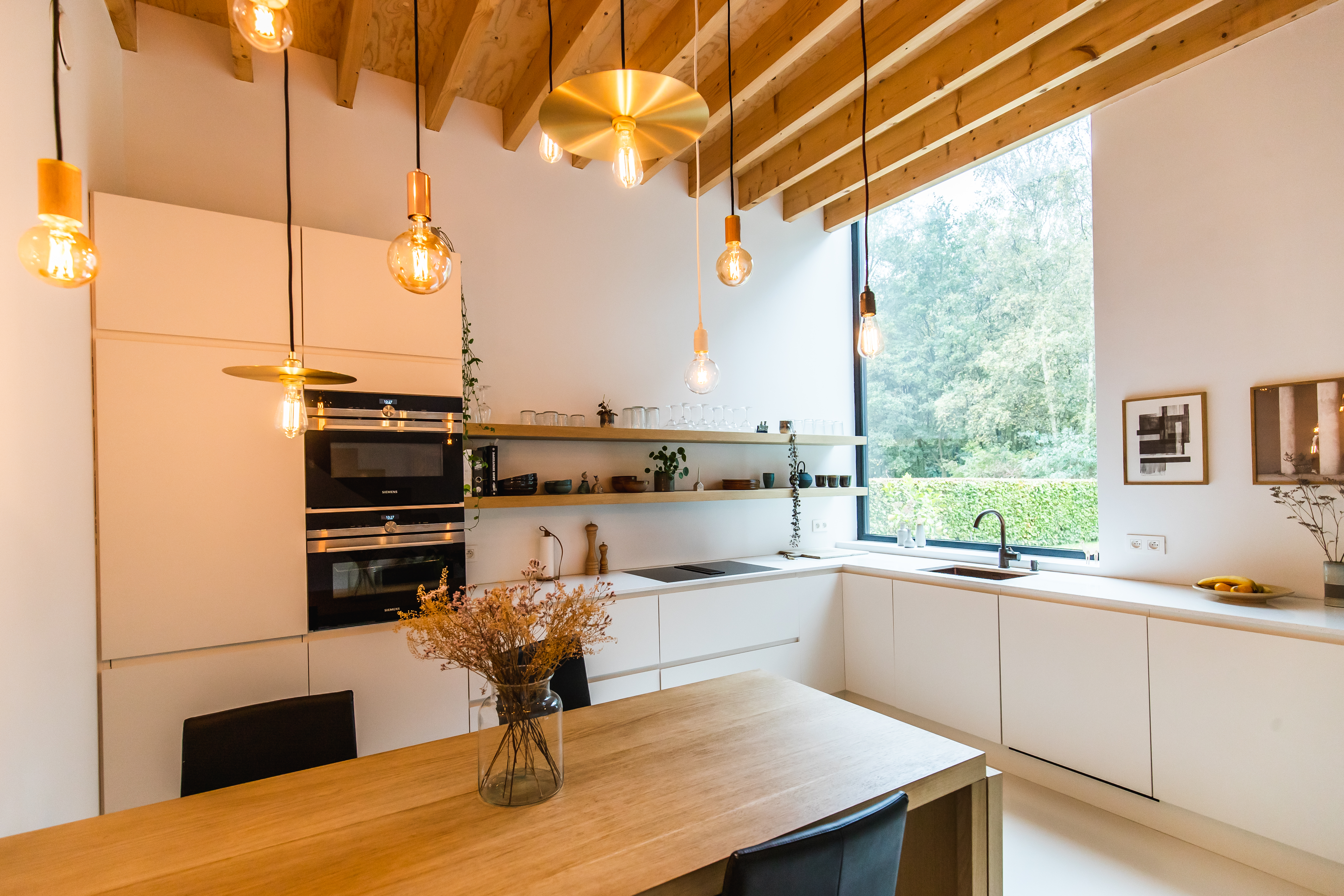 Moderne open keuken in een vrijstaande nieuwbouwwoning met witte inbouwkasten, houten plafond, eettafel en groot raam met zicht op de tuin.