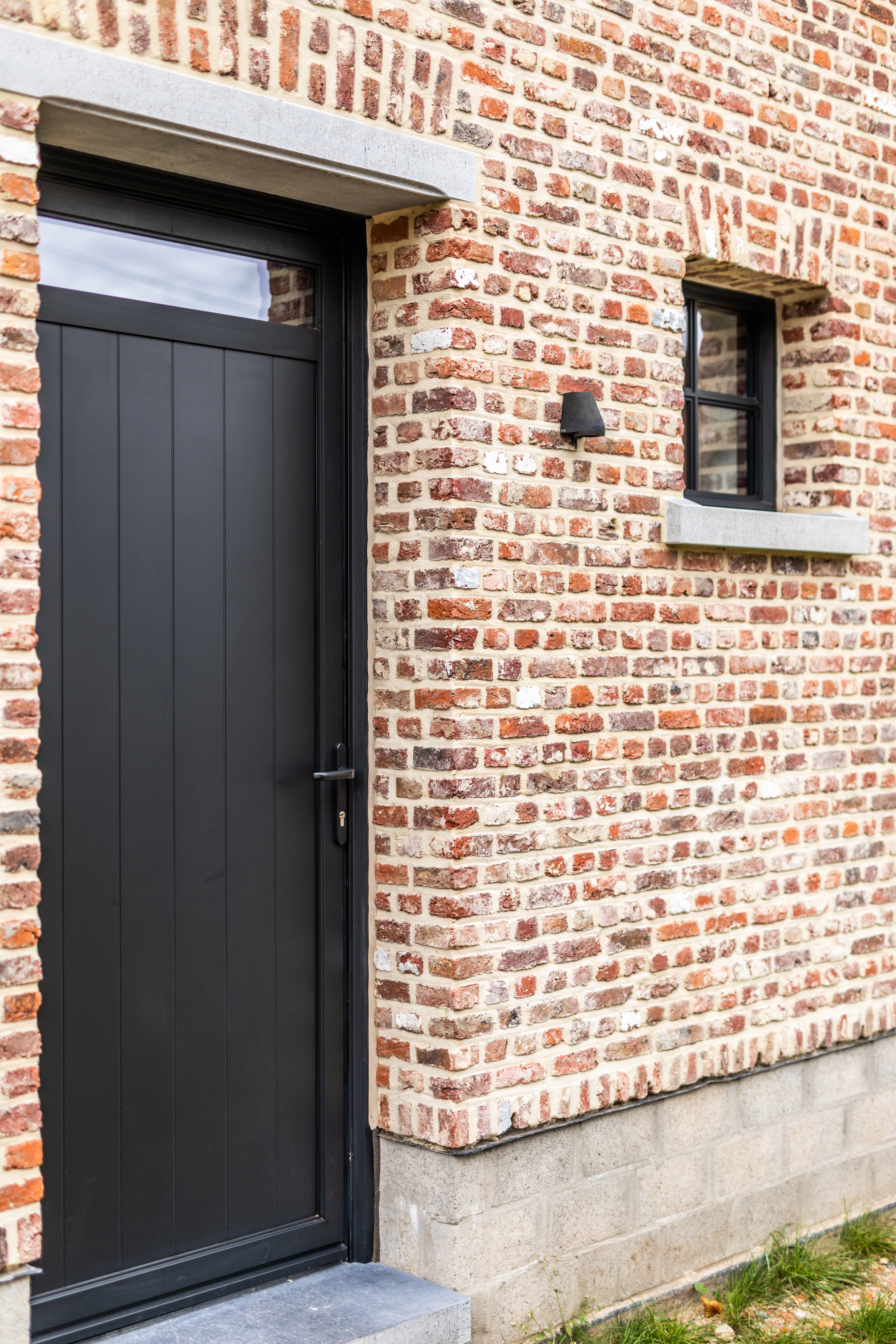 Zwarte ALU Steellook PLUS-buitendeur met bovenlicht in een warme, landelijke bakstenen gevel, naast een klein raam van een vrijstaande nieuwbouwwoning.