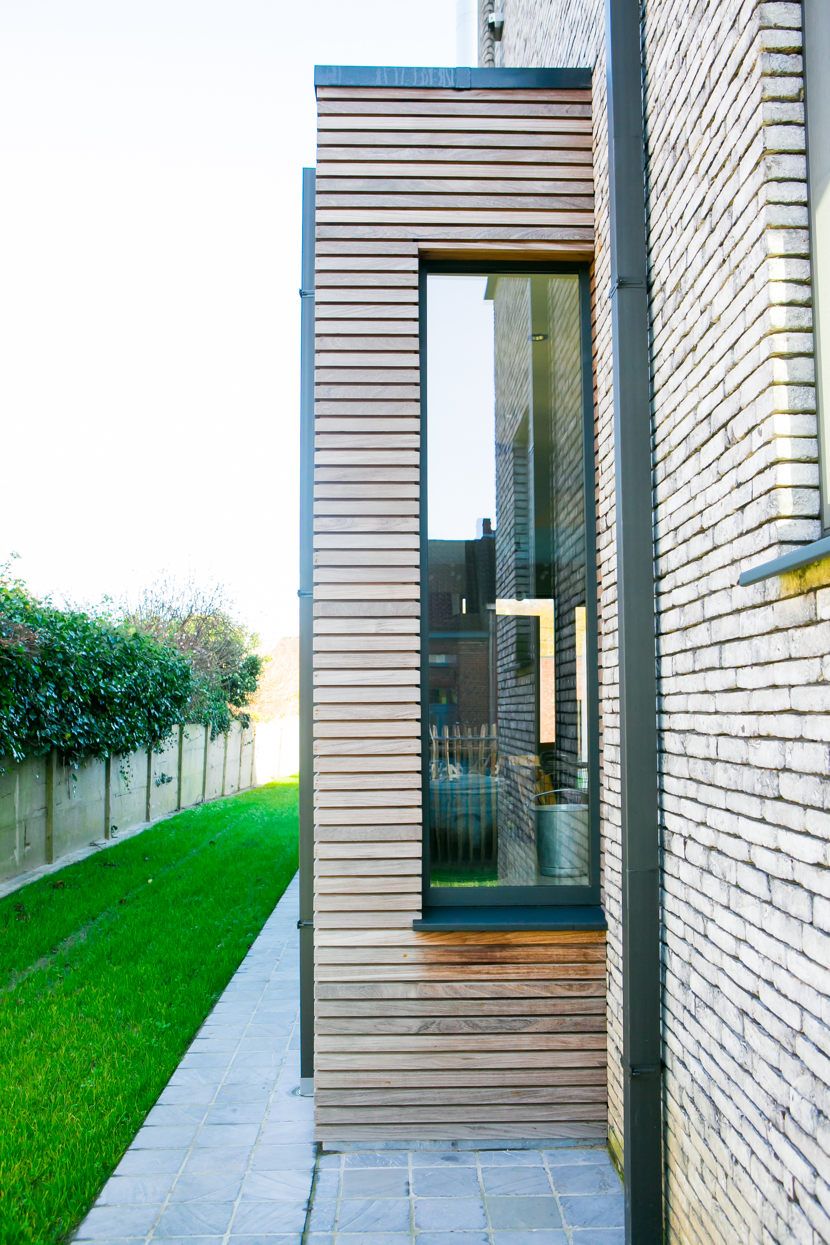 Modern vast raam met zwartgrijze raamprofielen, geïntegreerd in de houten gevel van een nieuwbouwwoning, naast een lichte bakstenen muur en een groene tuin.