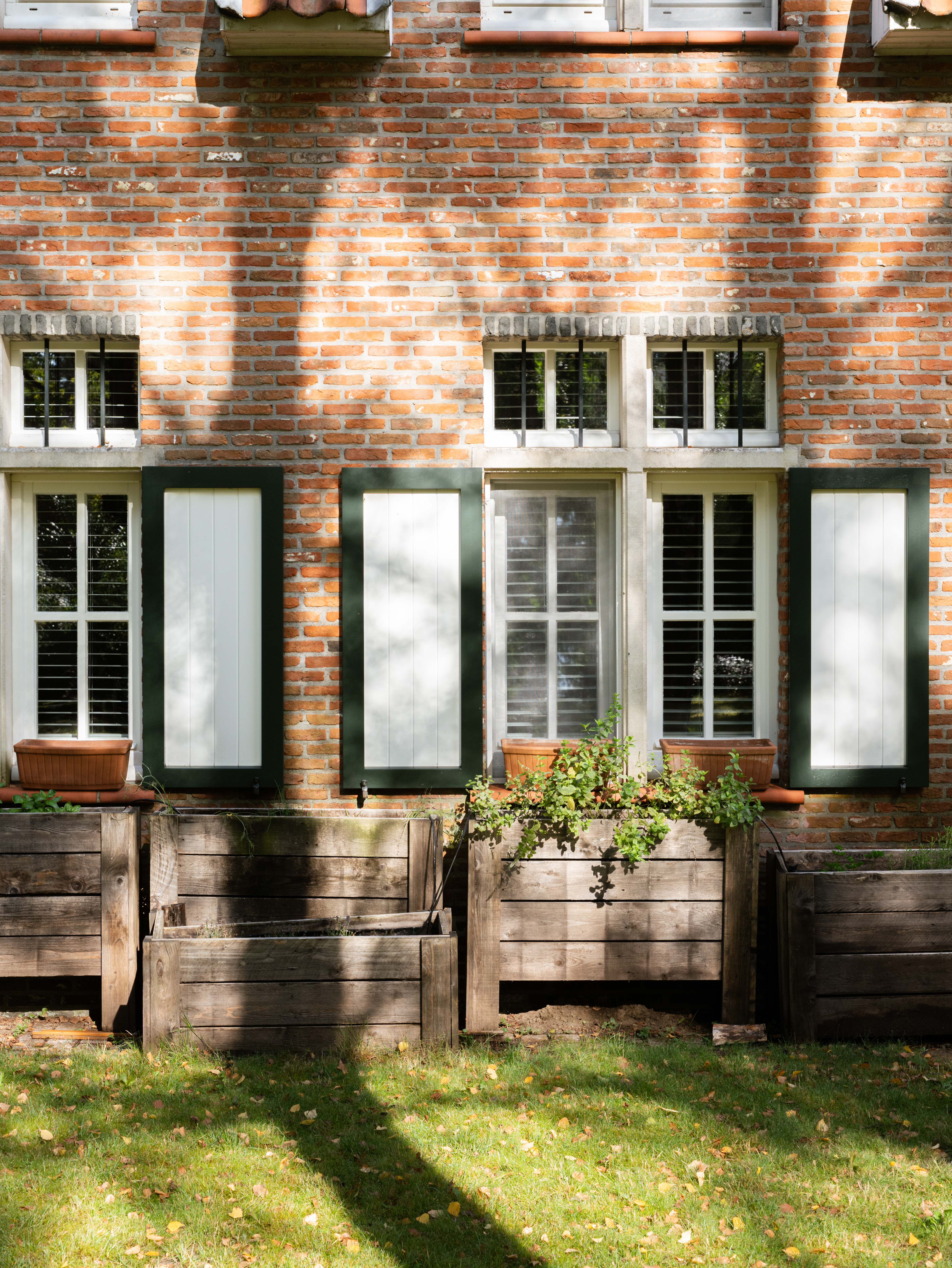 Warme, landelijk ogende bakstenen gevel met lichte ramen en donkere luiken. Houten plantenbakken staan voor de gerenoveerd ogende gevel van een vrijstaande woning.