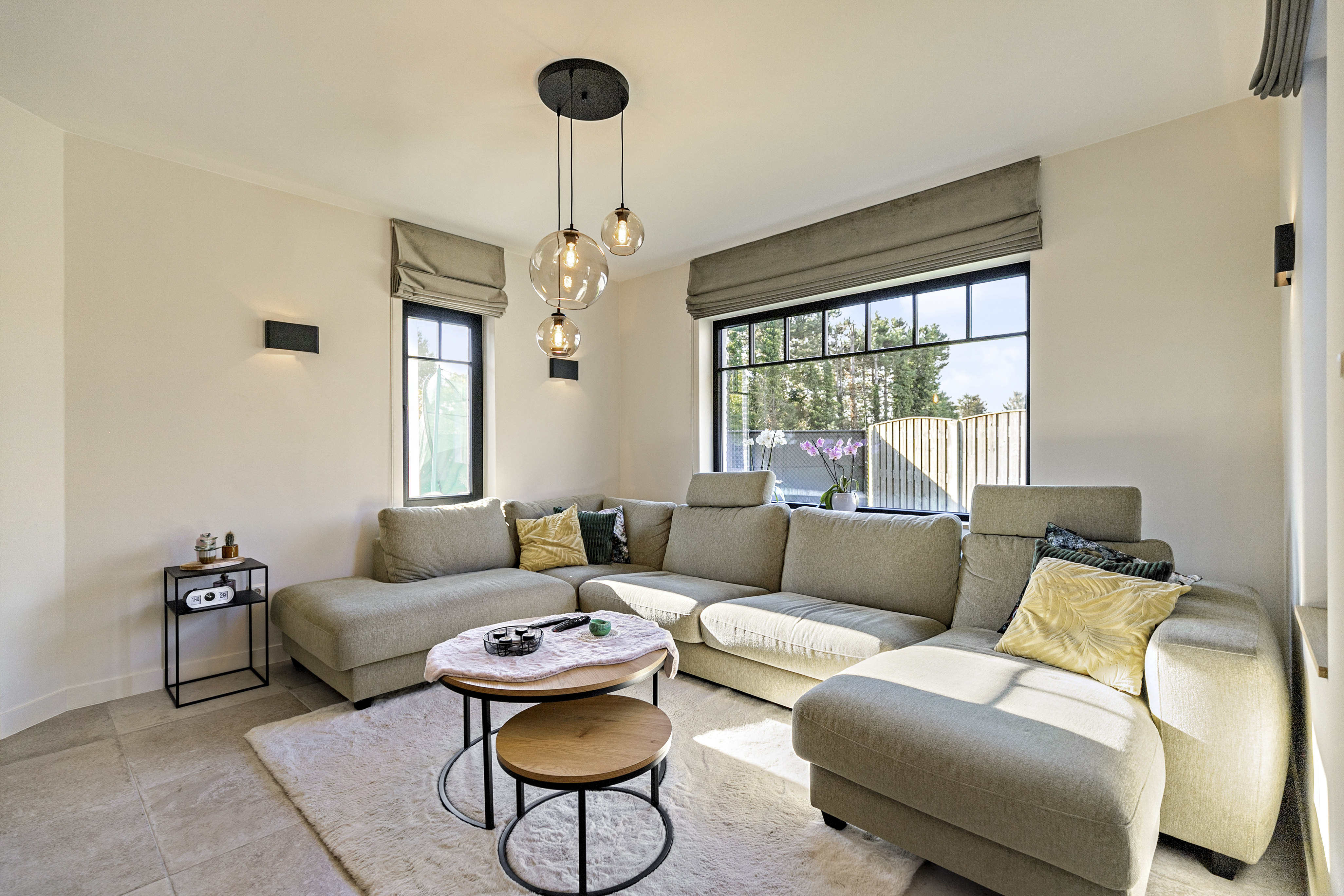 Lichte woonkamer met een matzwart draai-kipraam en vast raam, naast een hoeksofa, salontafels en zicht op de tuin.