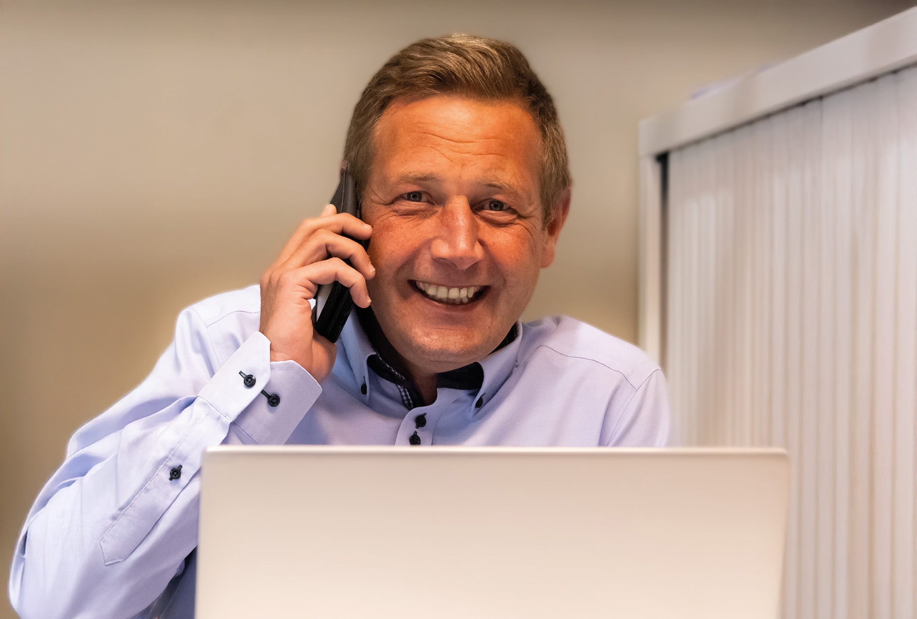 Een glimlachende man telefoneert achter een laptop in een kantoor met verticale lamellen.