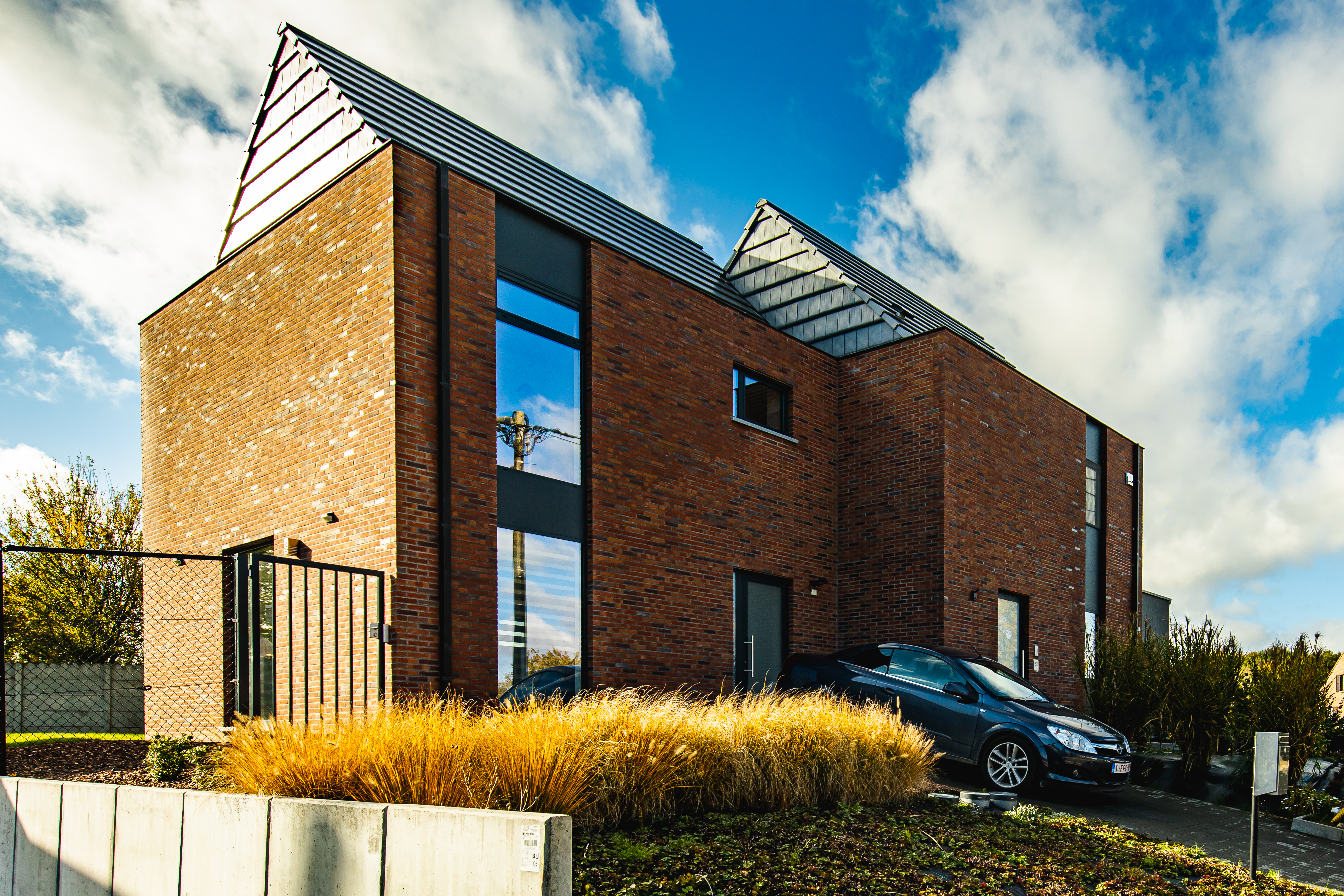 Vrijstaande nieuwbouwwoning in baksteen met antracietgrijze ramen en deuren, opvallende dakvormen, een tuin en een auto op de oprit.