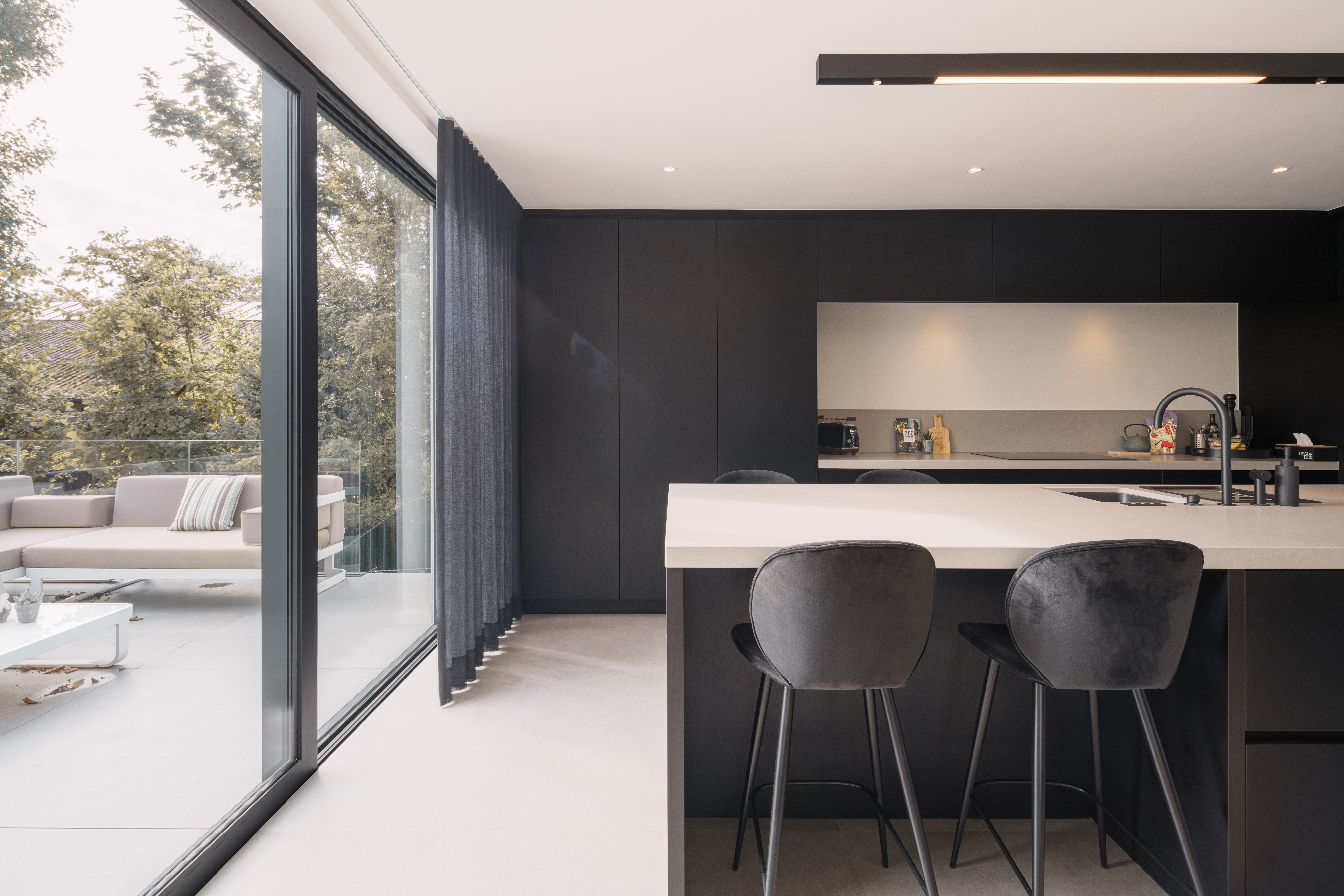 Modern nieuwbouwinterieur met een zwart aluminium schuifraam van het type ALU Visoglide Monorail, dat de keuken met het lichte terras verbindt.