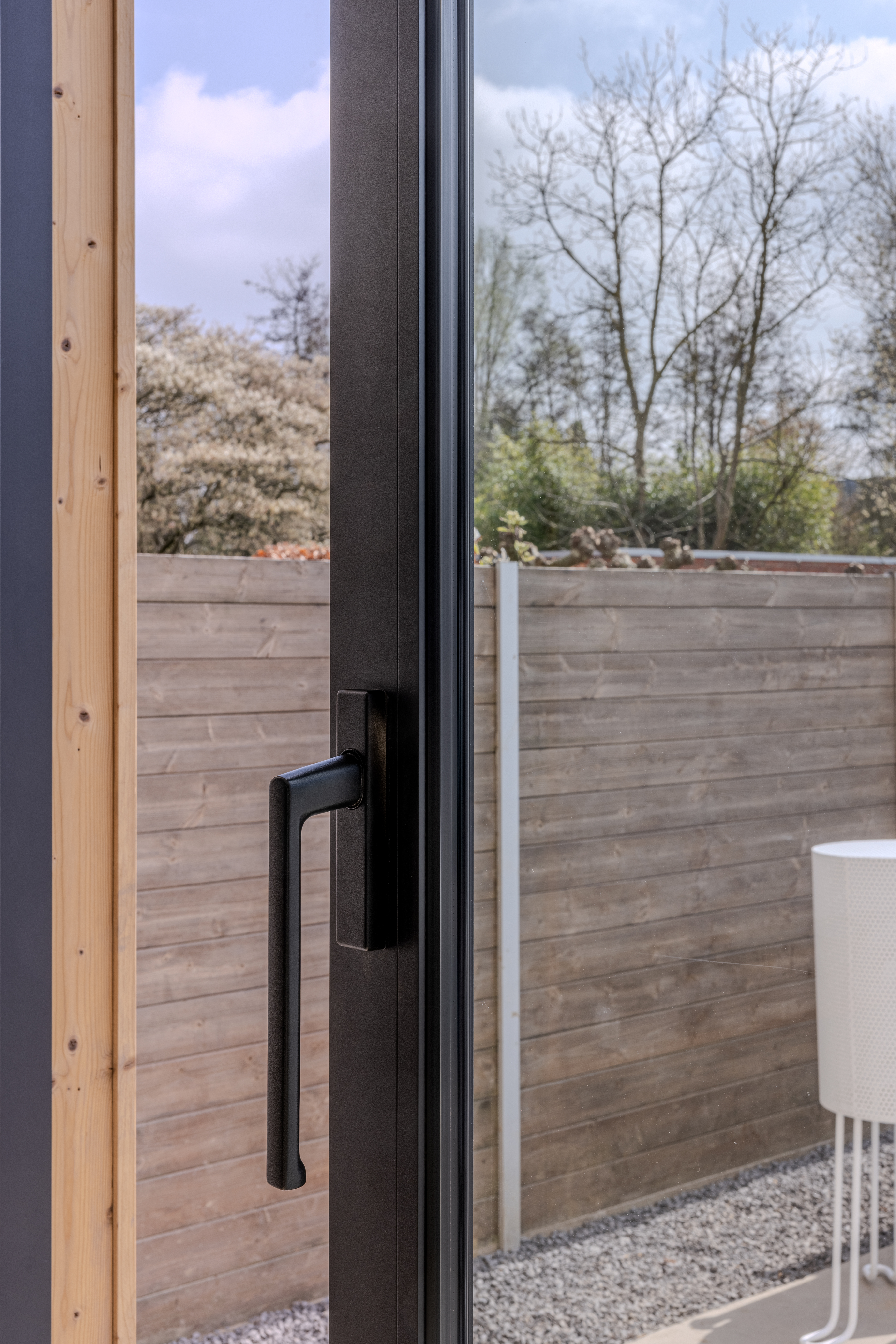 Close-up van een zwart schuifraam met lange handgreep naast een licht houten raamprofiel, met een houten afsluiting, grind en tuin op de achtergrond.