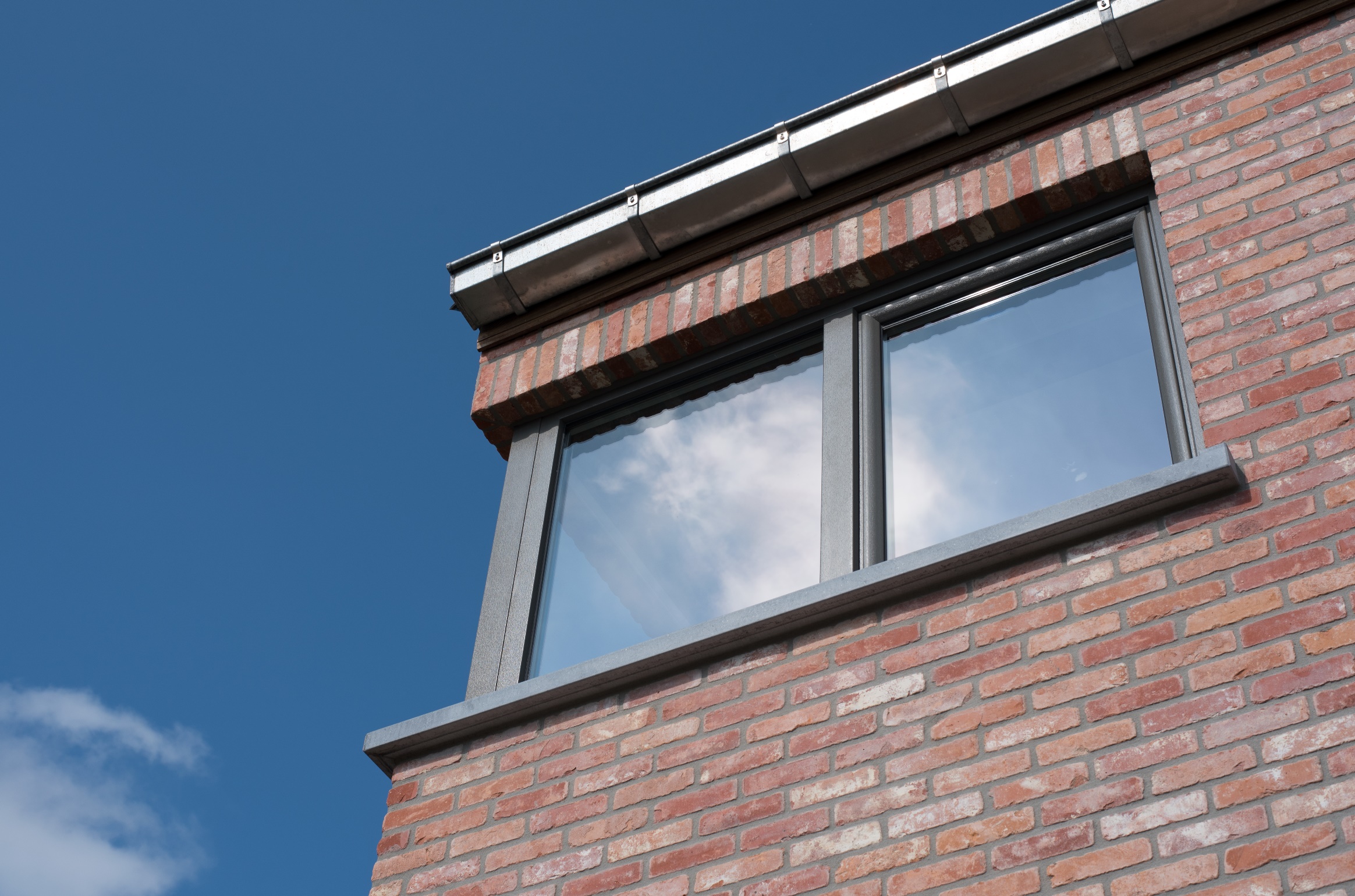 Kwartsgrijze NERF-raamprofielen rond een raam met twee verticale delen in de bakstenen gevel van een halfopen nieuwbouwwoning.
