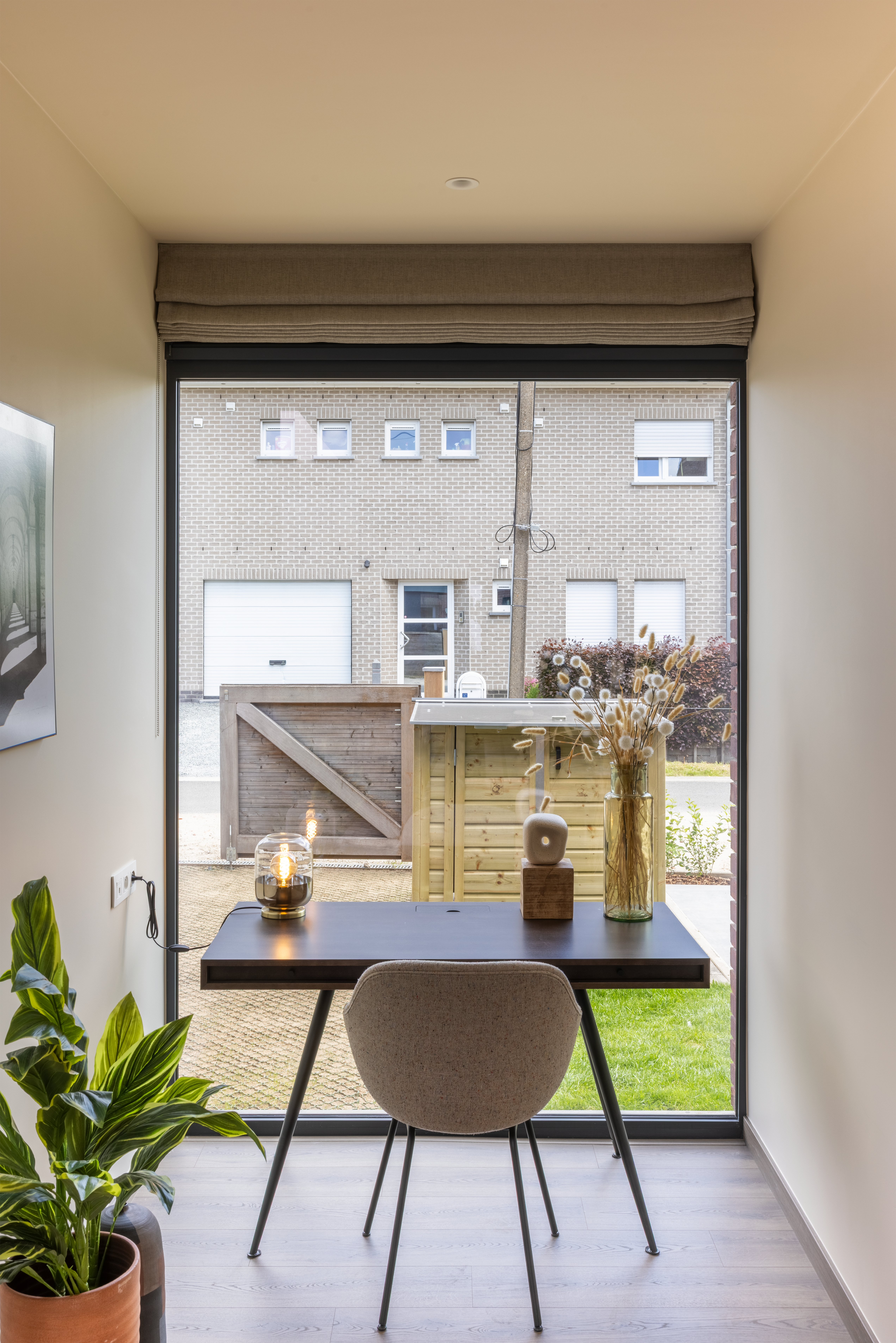 Groot vast raam met antracietgrijze raamprofielen in een gemoedelijk, tijdloos interieur met tafel en stoel, met zicht op een gesloten bebouwing.