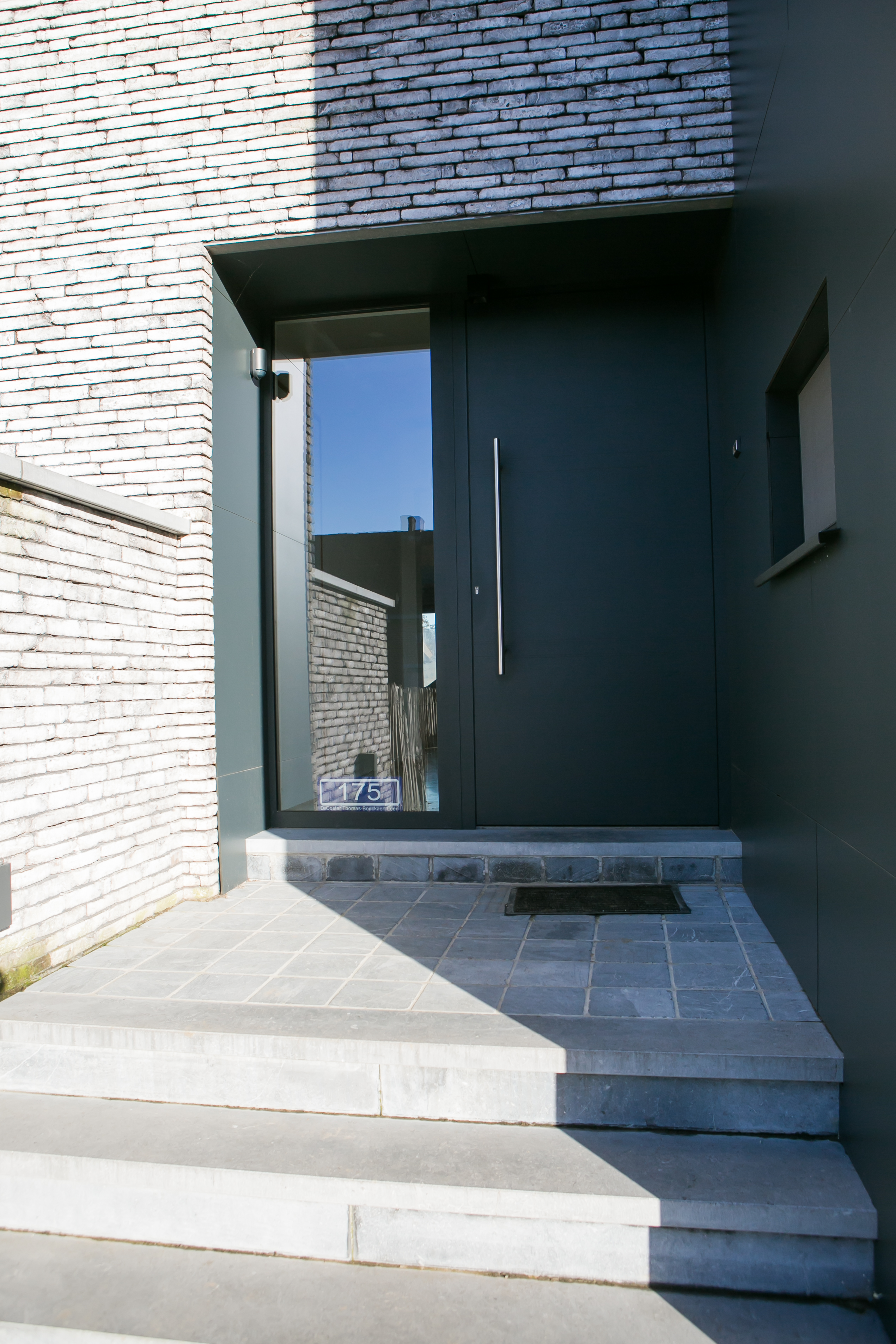 Moderne inkom van een nieuwbouwwoning met zwartgrijze aluminium voordeur van ALU Koncept, smal zijlicht en brede stenen trap.