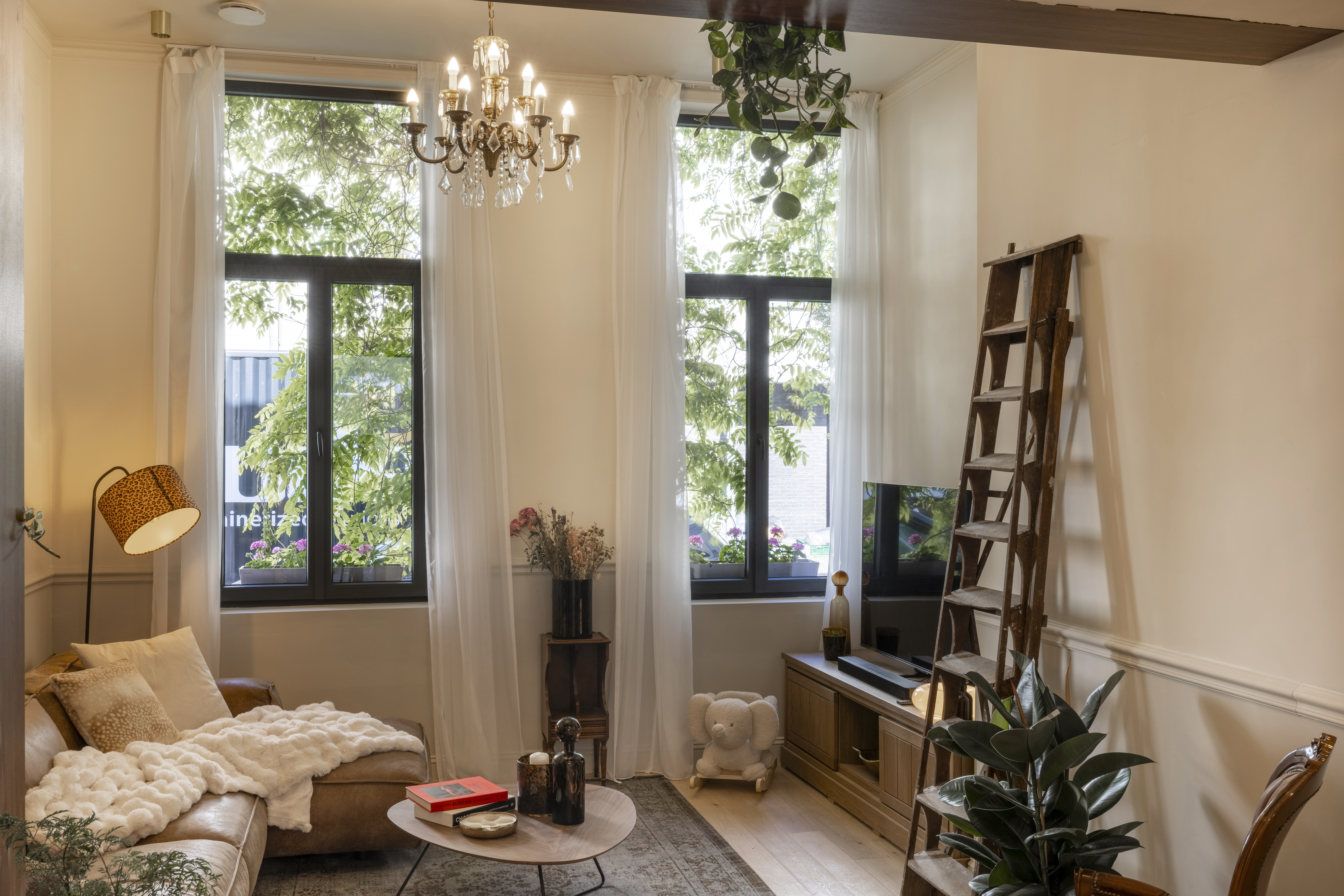 Lichte woonkamer met twee hoge ramen in mat brons, gecombineerd met bovenlicht, vast raam en draai-kipraam, naast een ladder en zitmeubels.