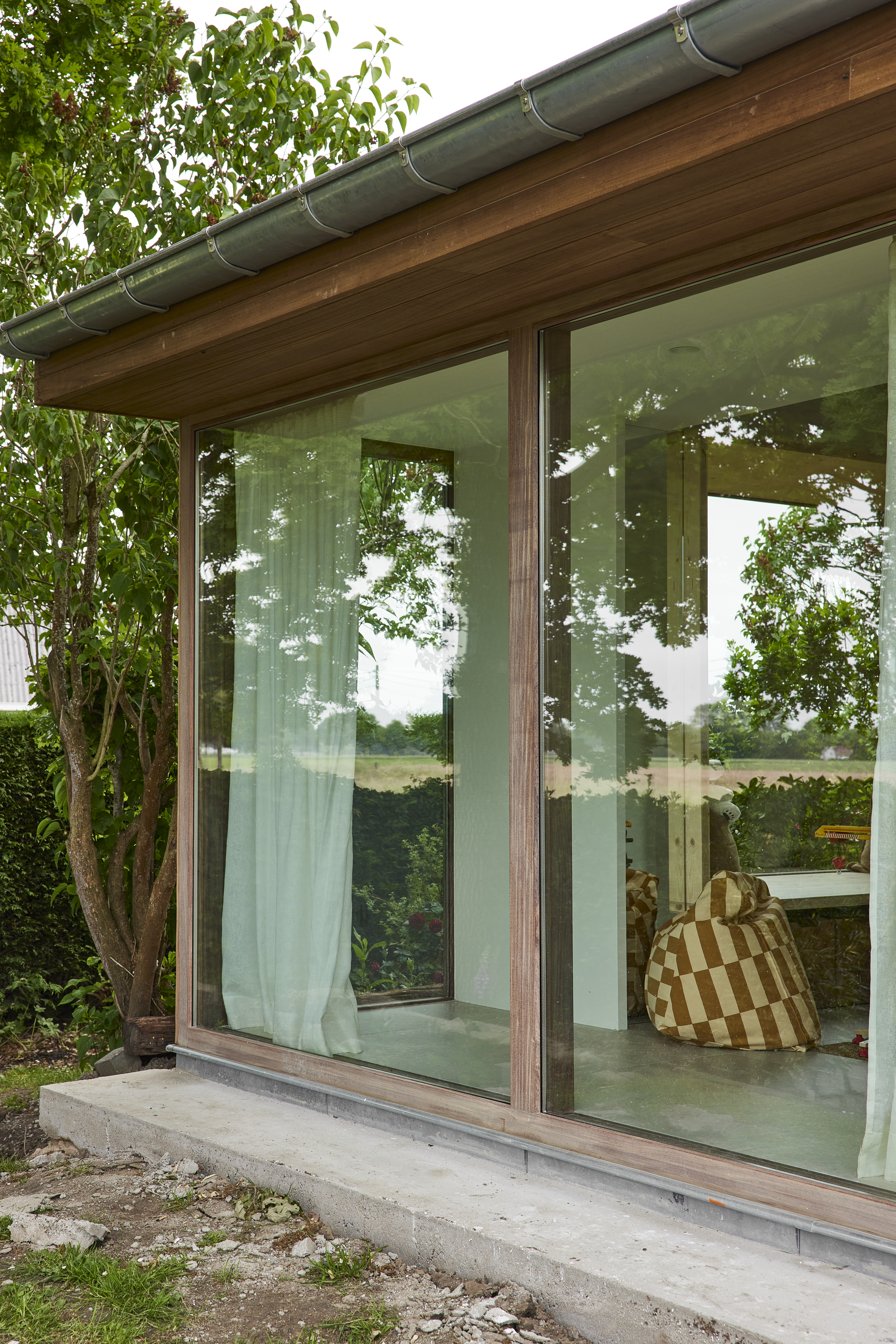 Grote glazen gevel met houten raamprofielen in Afromosia, afgewerkt met Rubio Natural, naast een tuin en onder een brede houten dakoverstek.