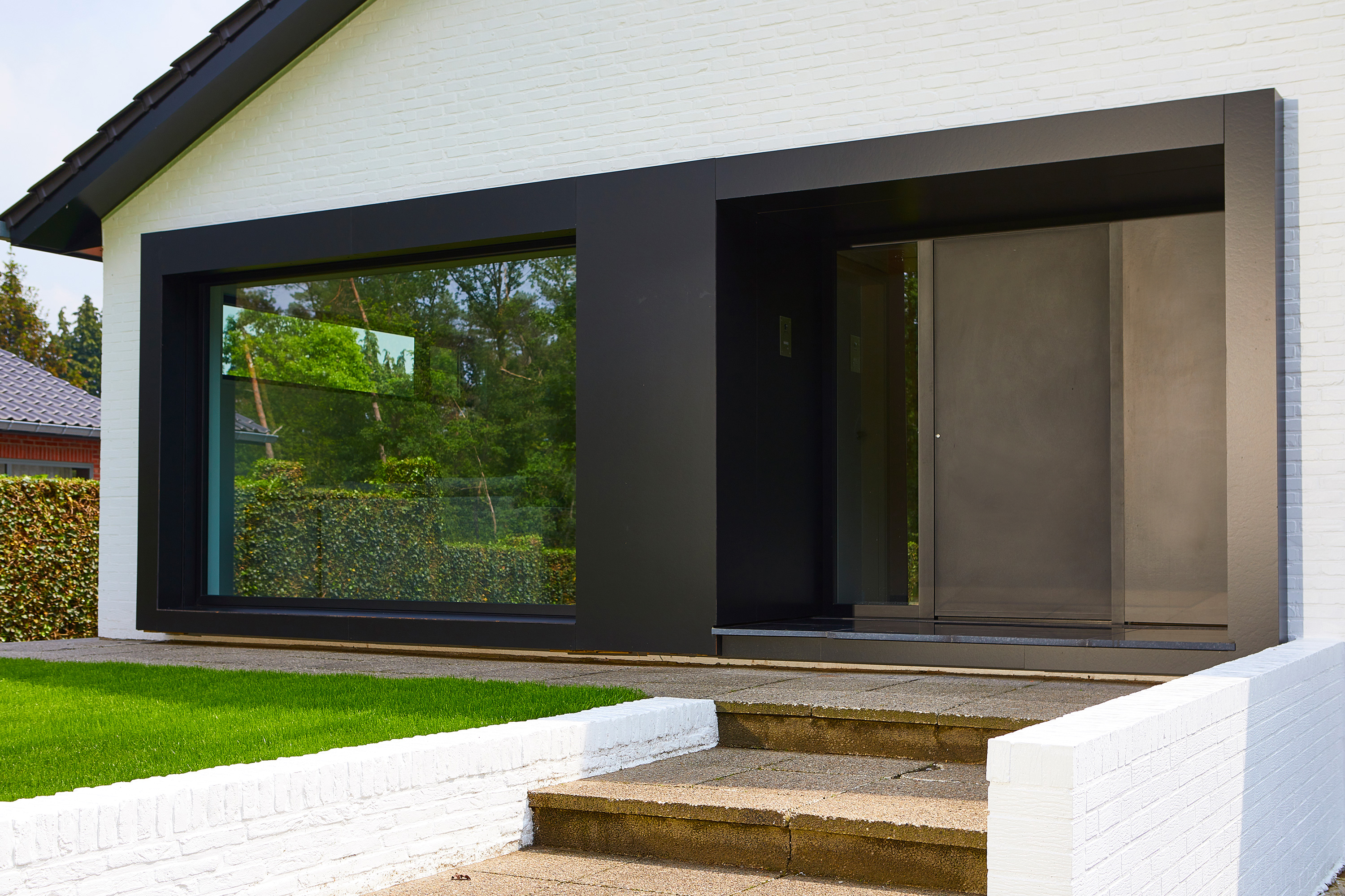 Moderne gerenoveerde gevel van een vrijstaande woning met zwarte steellookramen, een inkomdeur met zijlicht, witte bakstenen en toegangstreden.