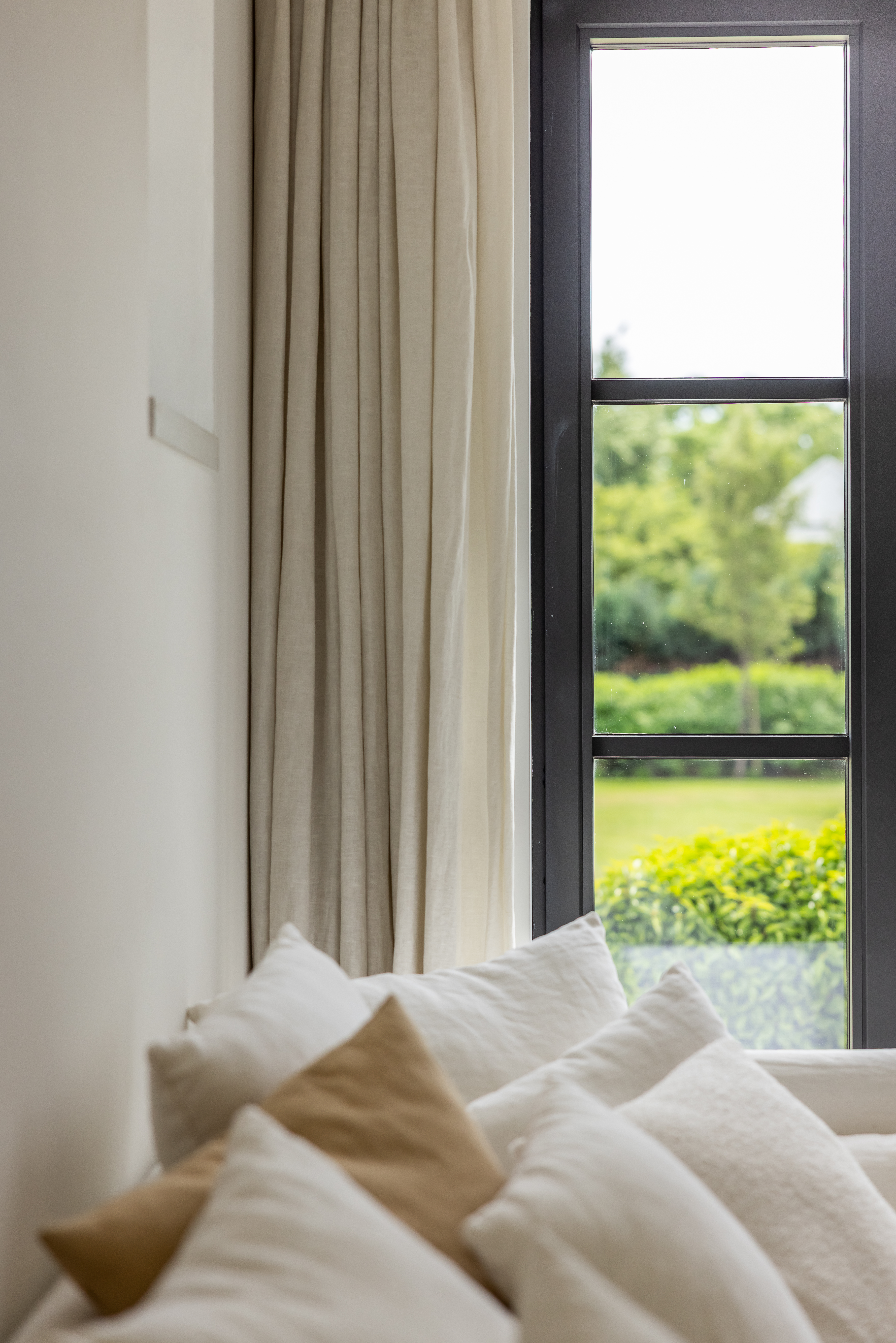 Smalle zwarte Steel-Look PLUS-ramen in een warm landelijk interieur, omlijst door beige gordijnen en kussens, met zicht op het groen.