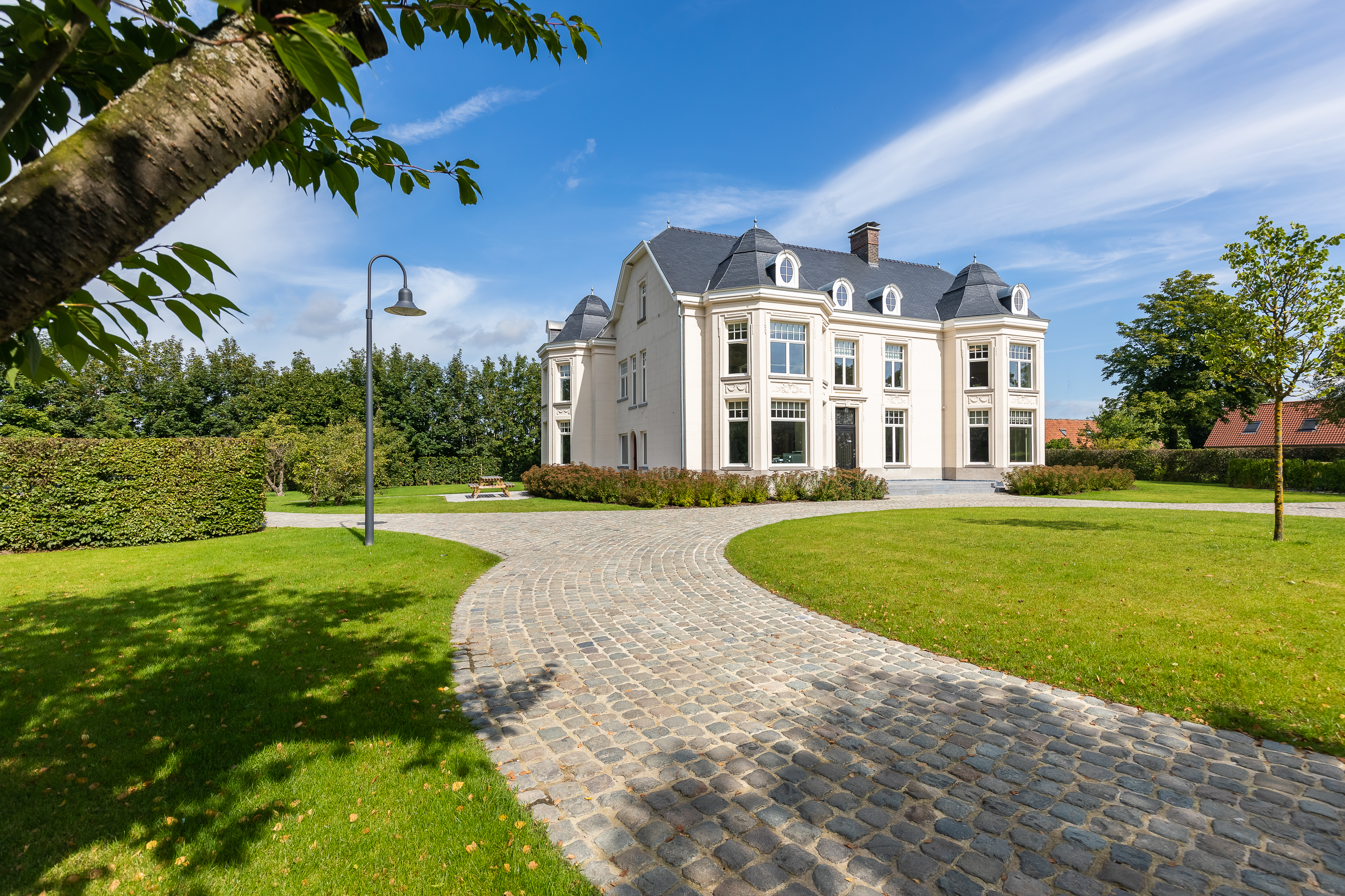 Witte, gerenoveerde vrijstaande woning in verfijnde klassieke stijl, met hoge ramen, mansardedak, verzorgd gazon en een gebogen oprit in kasseien.