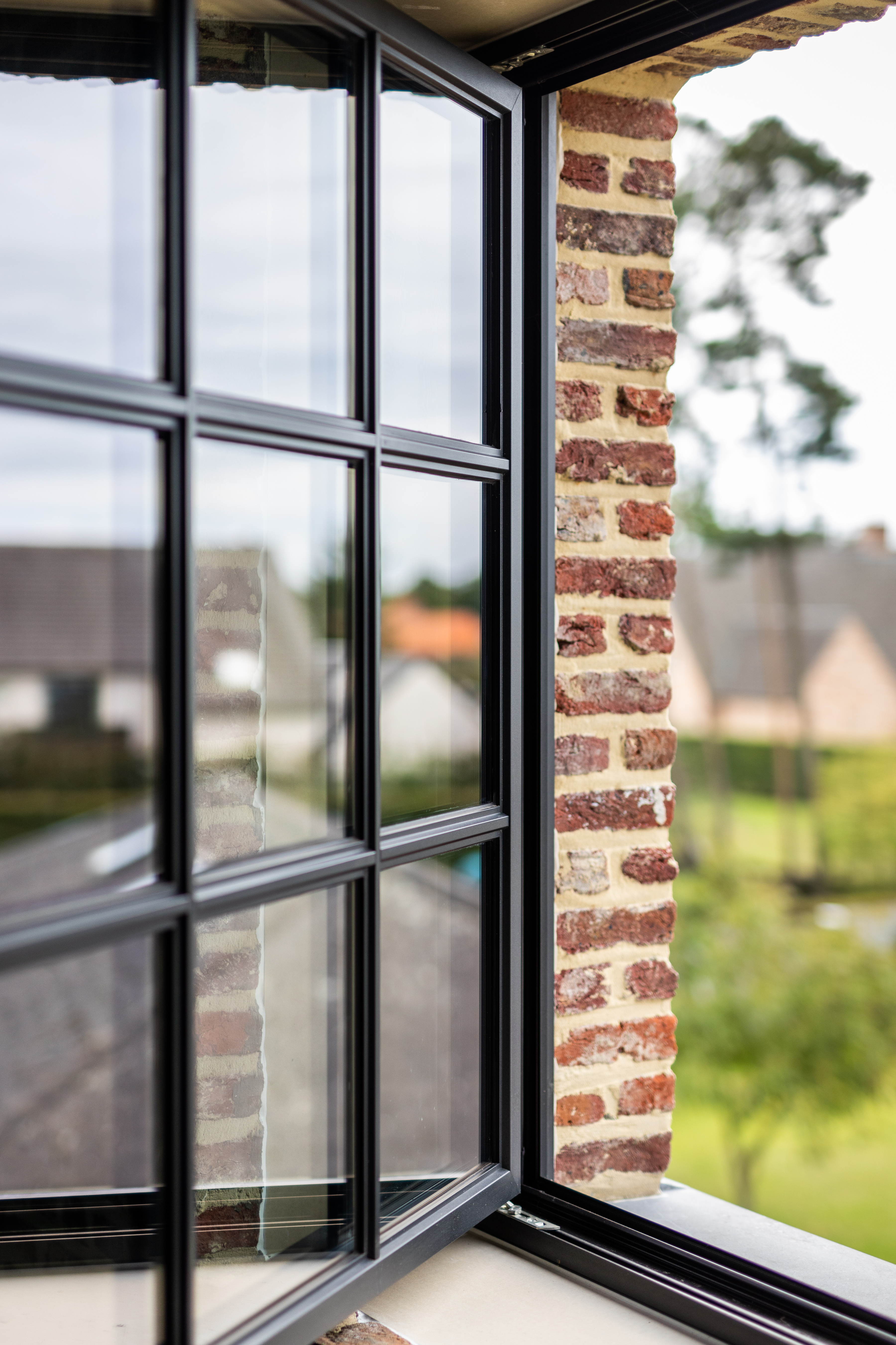 Zwart steellook draai-kipraam in een warm landelijke bakstenen gevel van een open bebouwing, met slanke profielen en zicht op de groene omgeving.