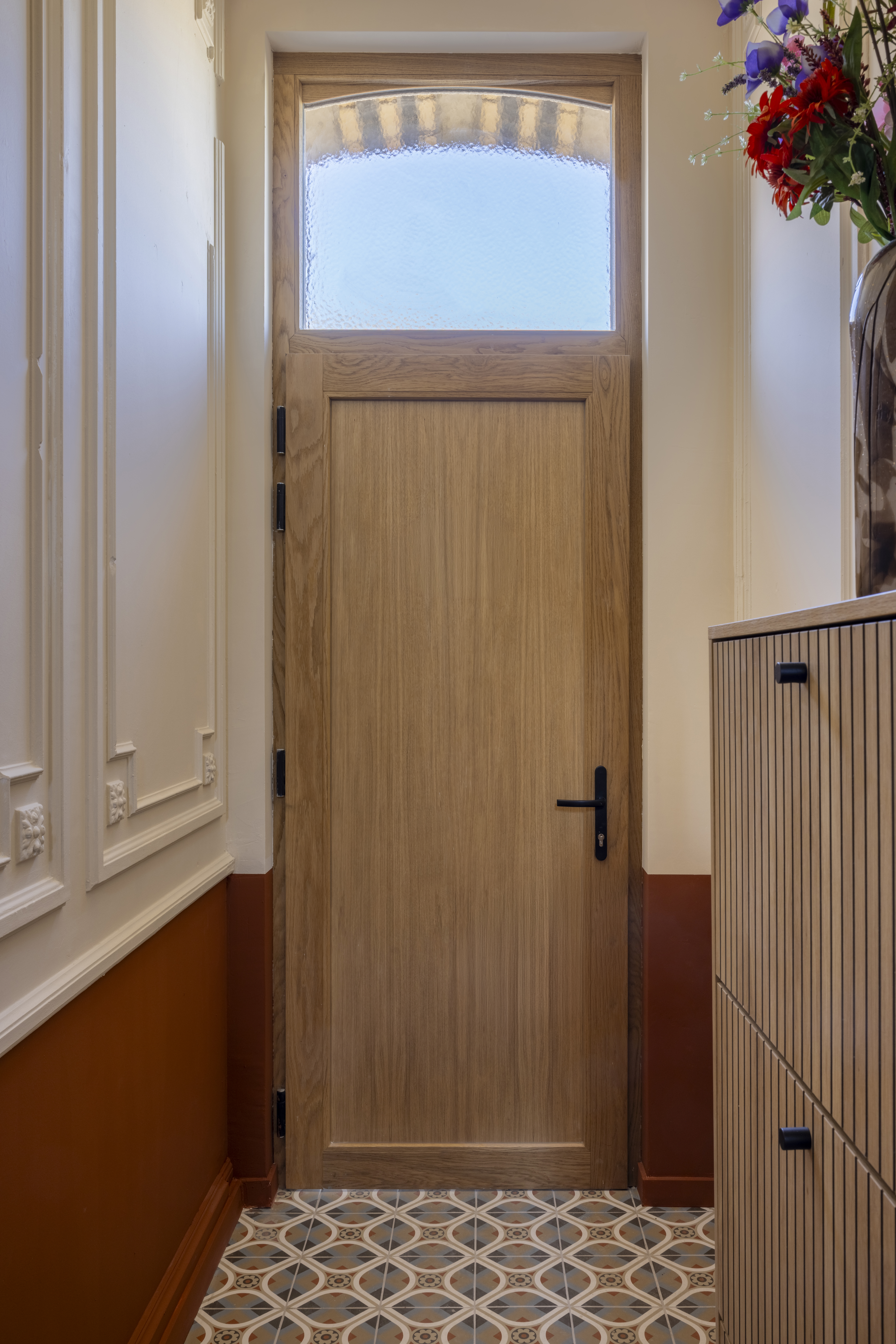 Eikenhouten deur met bovenlicht in een smalle inkomhal, met een decoratieve vloer en houten wandbekleding.