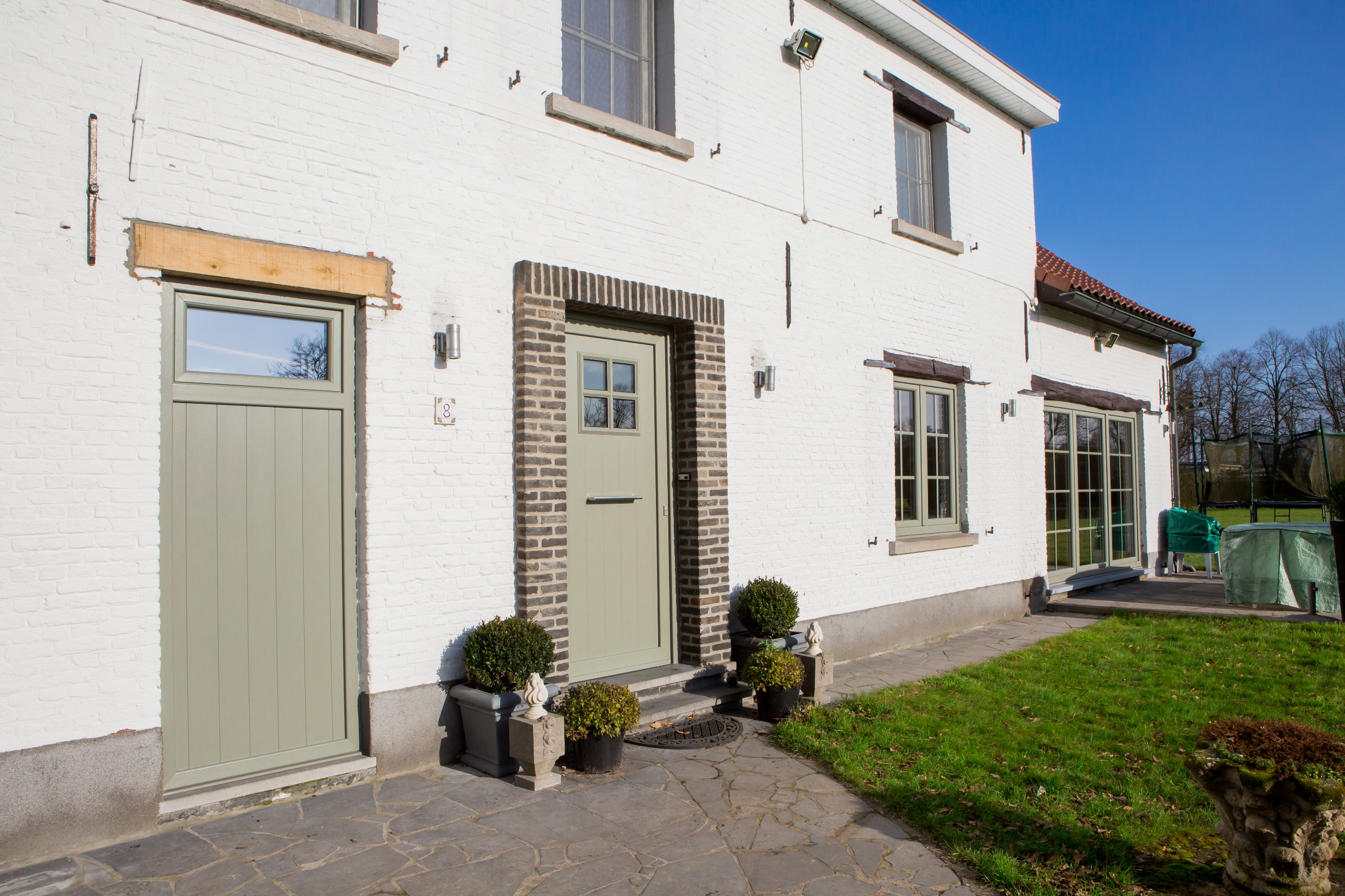Gerenoveerde open bebouwing met betongrijze NERF-panelen, lichte gevel, grijsgroene ramen en deuren en een kleine tuin.