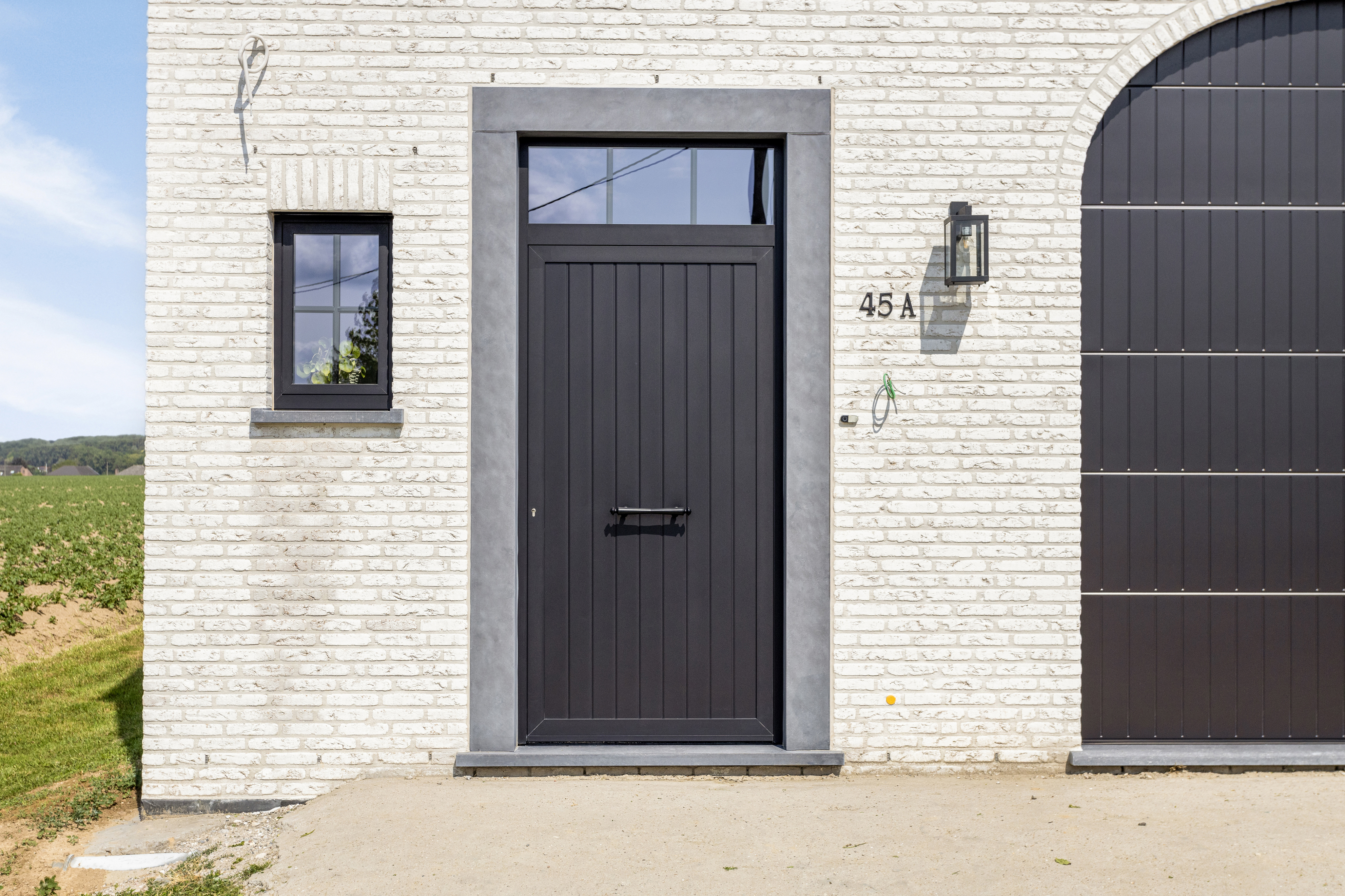 Signaalzwarte paneeldeur met bovenlicht in de lichte bakstenen gevel van een landelijke vrijstaande nieuwbouwwoning, naast een grote donkere garagepoort en een open landschap.