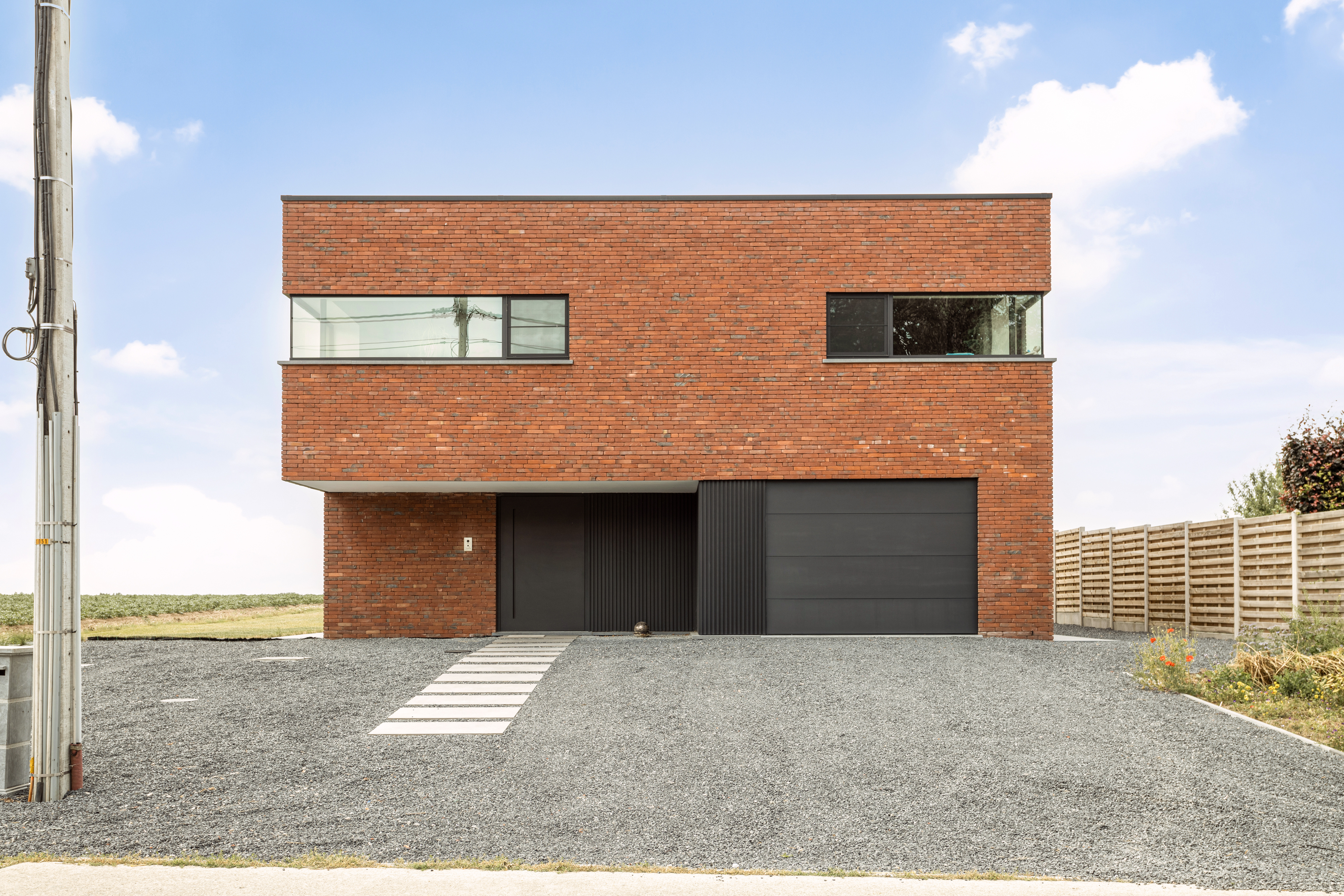 Moderne nieuwbouwwoning in open bebouwing met rode bakstenen gevel, zwarte garagepoort, hoekraam en vast raam, aan een grindoprit met open zicht op het landschap.