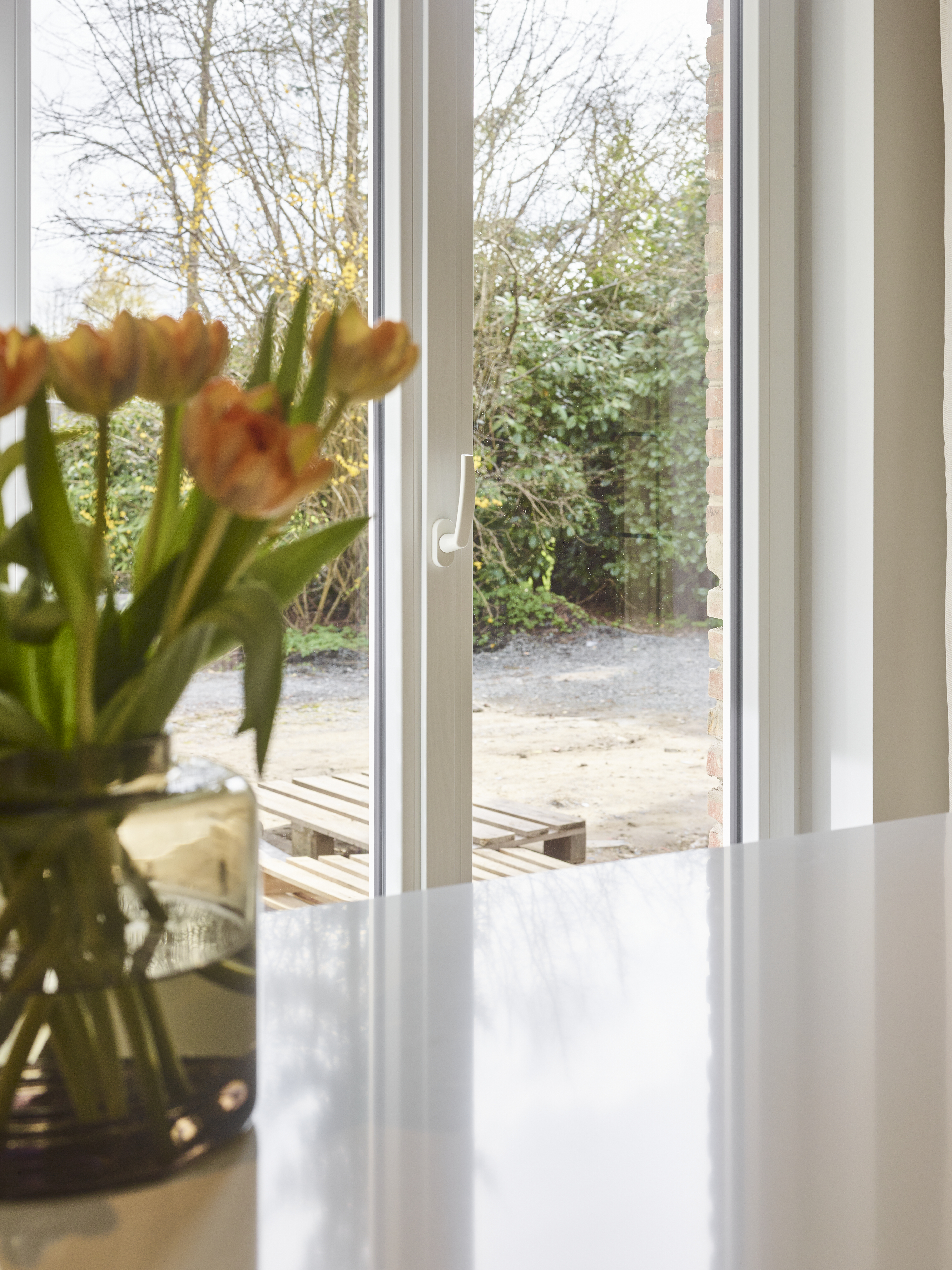 Wit dubbel draai-kipraam met slanke raamprofielen, uitzicht op de tuin en een vaas met tulpen in een licht interieur.