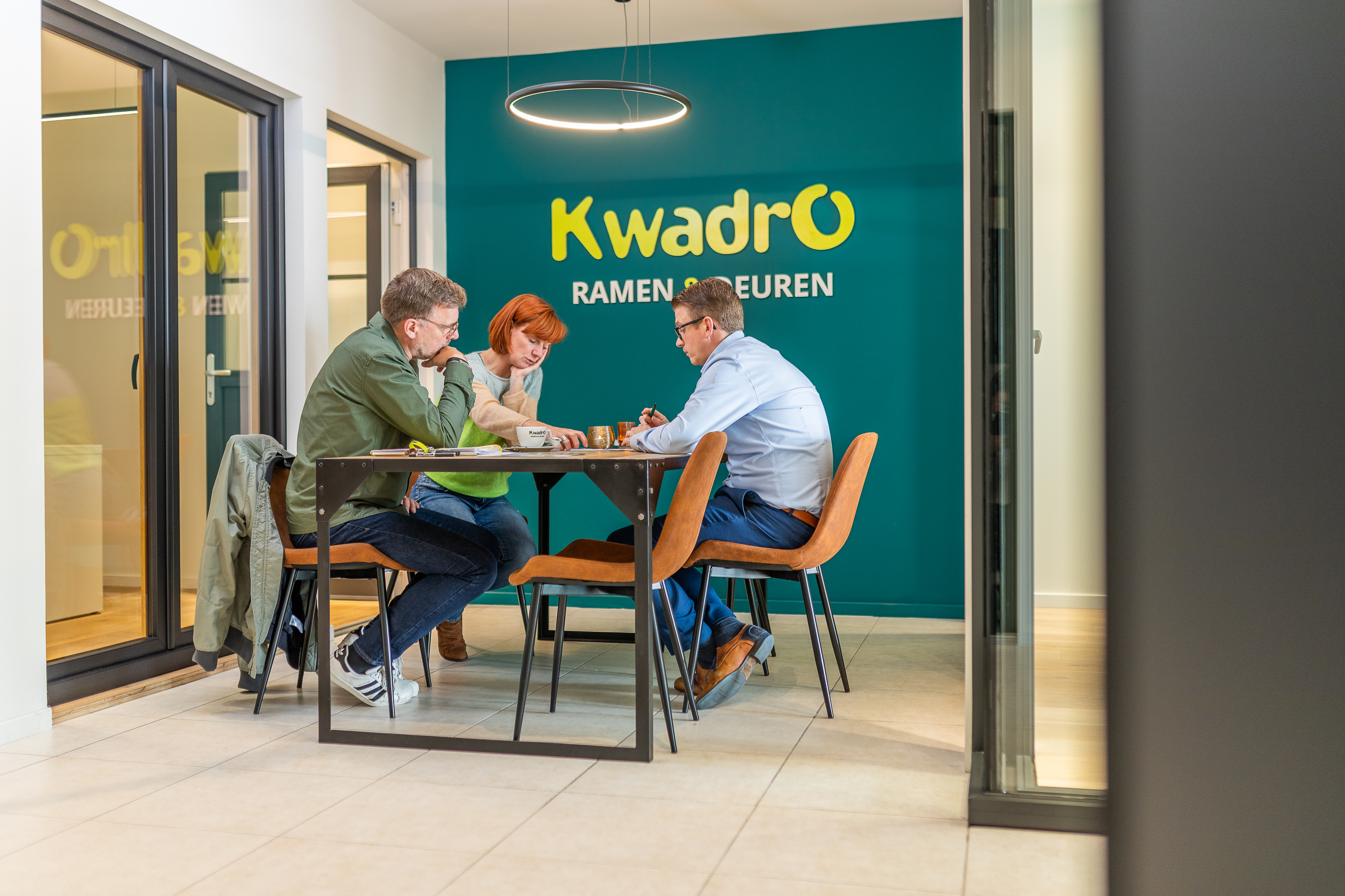 Drie personen bespreken samen iets aan een tafel in een showroom met ramen en deuren, met het Kwadro-logo op de achterwand.