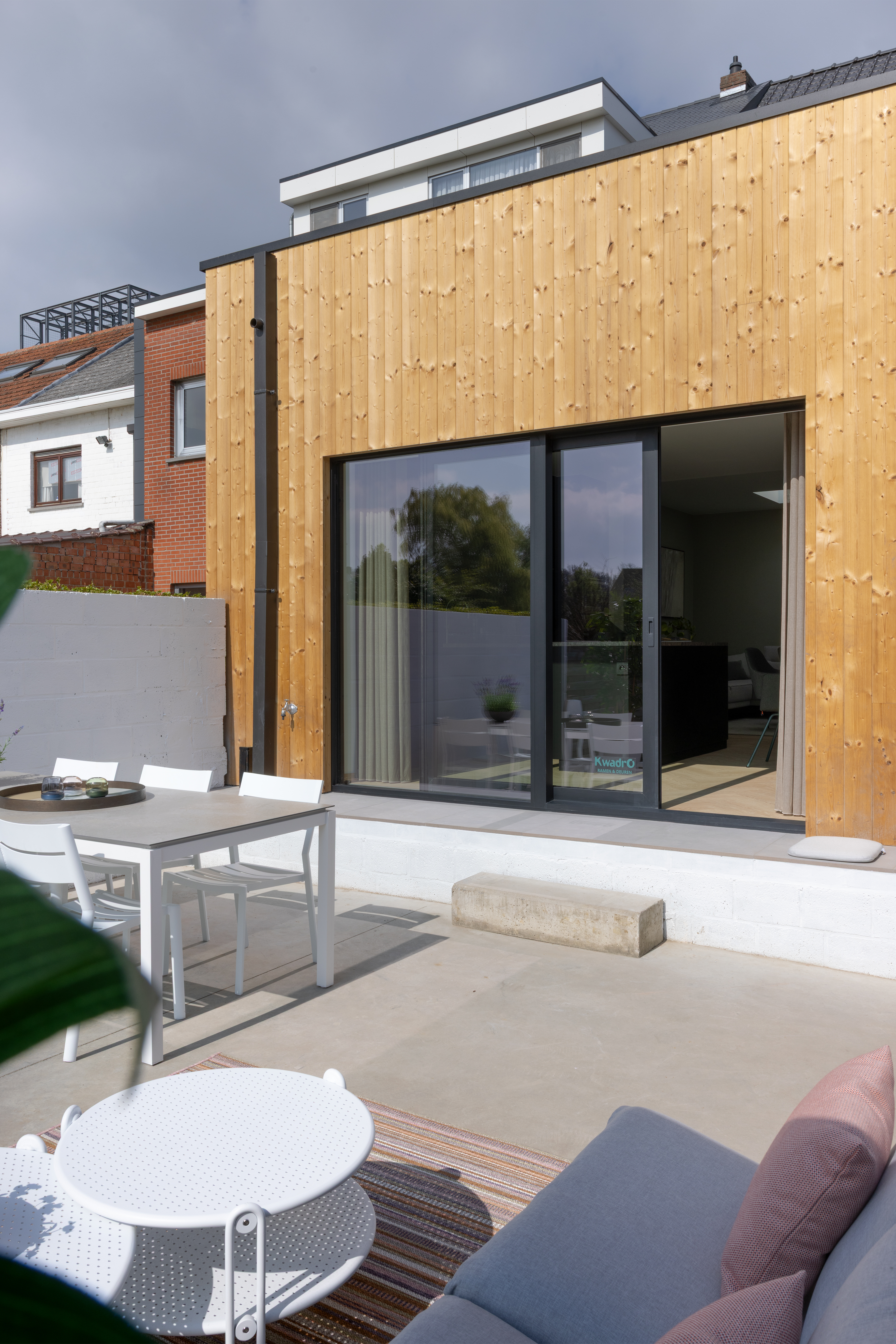 Moderne houten gevel met een groot zwart schuifraam naar een terras met tuinmeubelen en een gedeeltelijk zichtbare leefruimte.