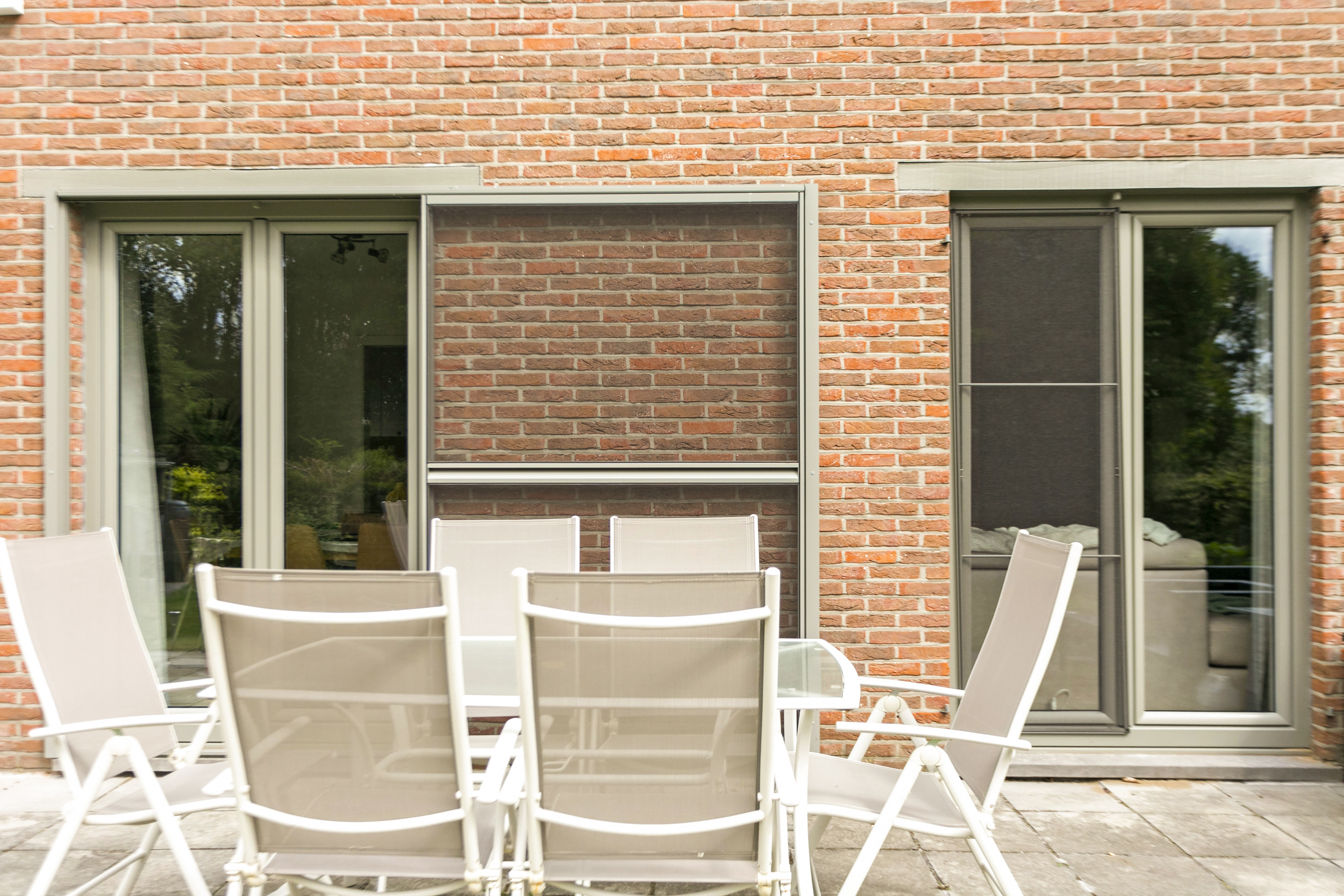Gerestaureerde bakstenen gevel van een open bebouwing met betongrijze dubbele draai-kipramen, vliegenramen en tuinmeubelen op het terras.