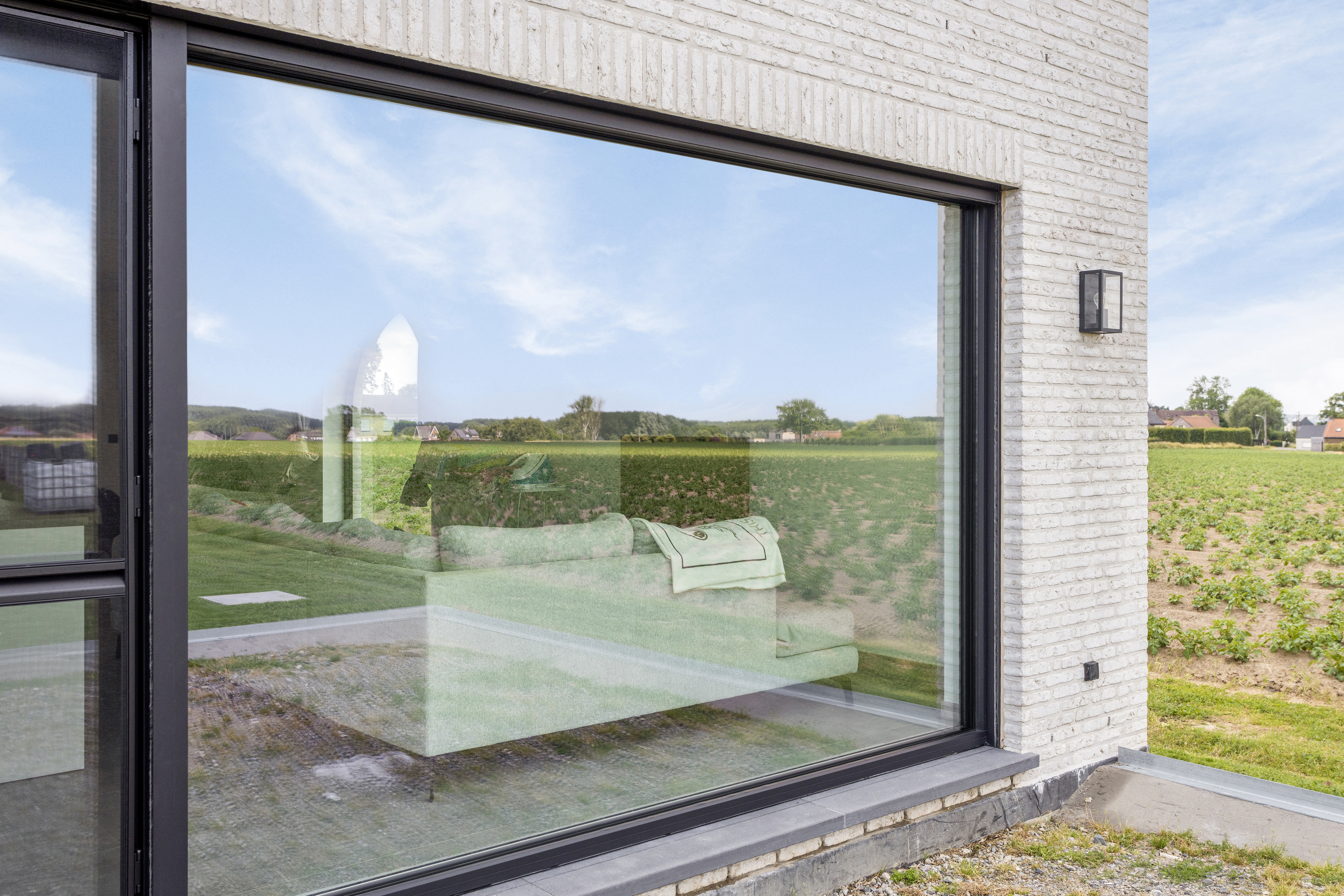 Groot vast raam met signaalzwarte raamprofielen in de lichte bakstenen gevel van een nieuwbouwwoning, met uitzicht op het landelijke landschap.