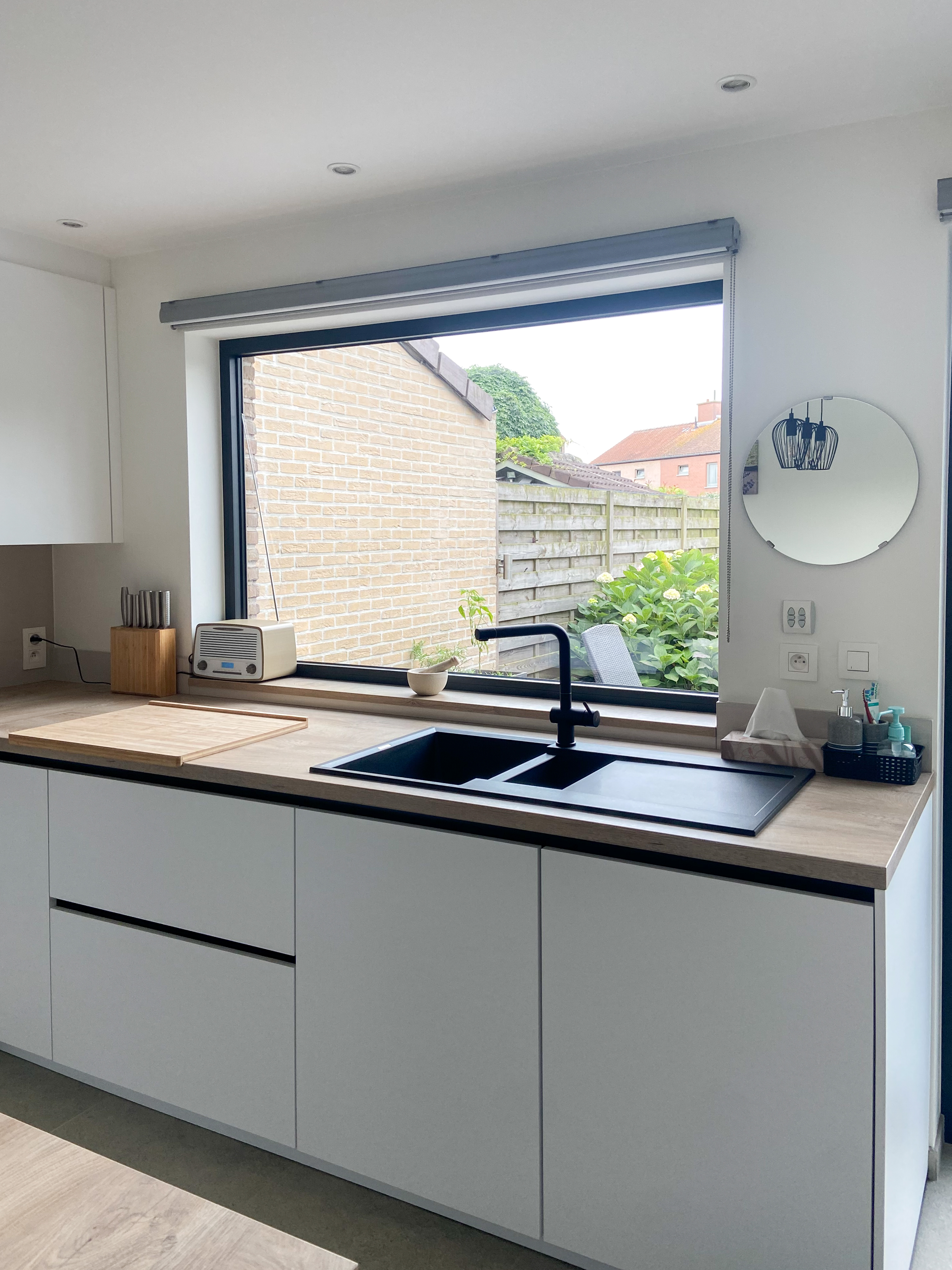 Gerenoveerde, tijdloze keuken in een vrijstaande woning met lichtgrijze kasten, houten werkblad, spoelbak en groot raam met zicht op de tuin.
