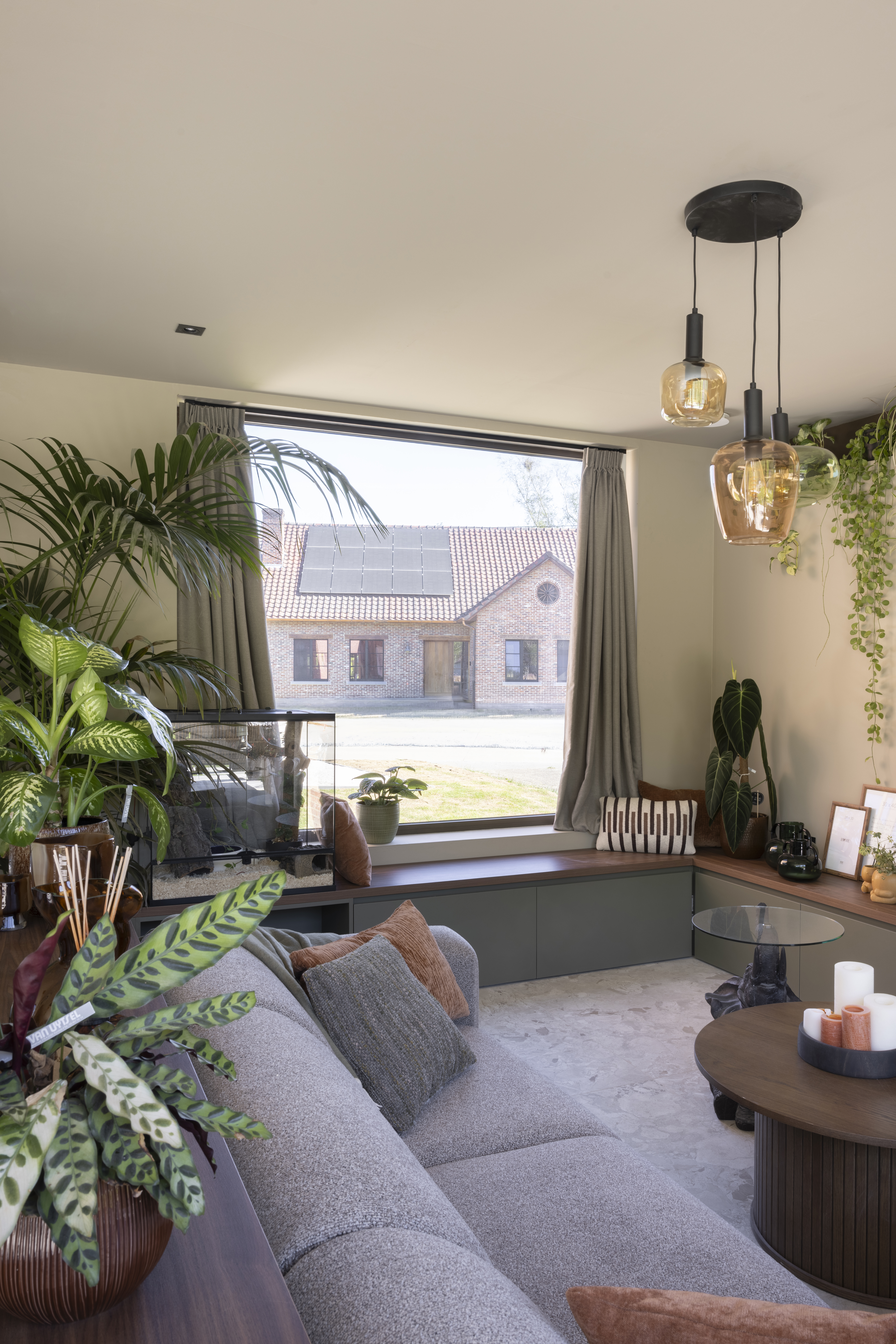 Lichte woonkamer met een groot vast raam met bronskleurig geanodiseerd raamprofiel, omringd door planten, een sofa en hanglampen.