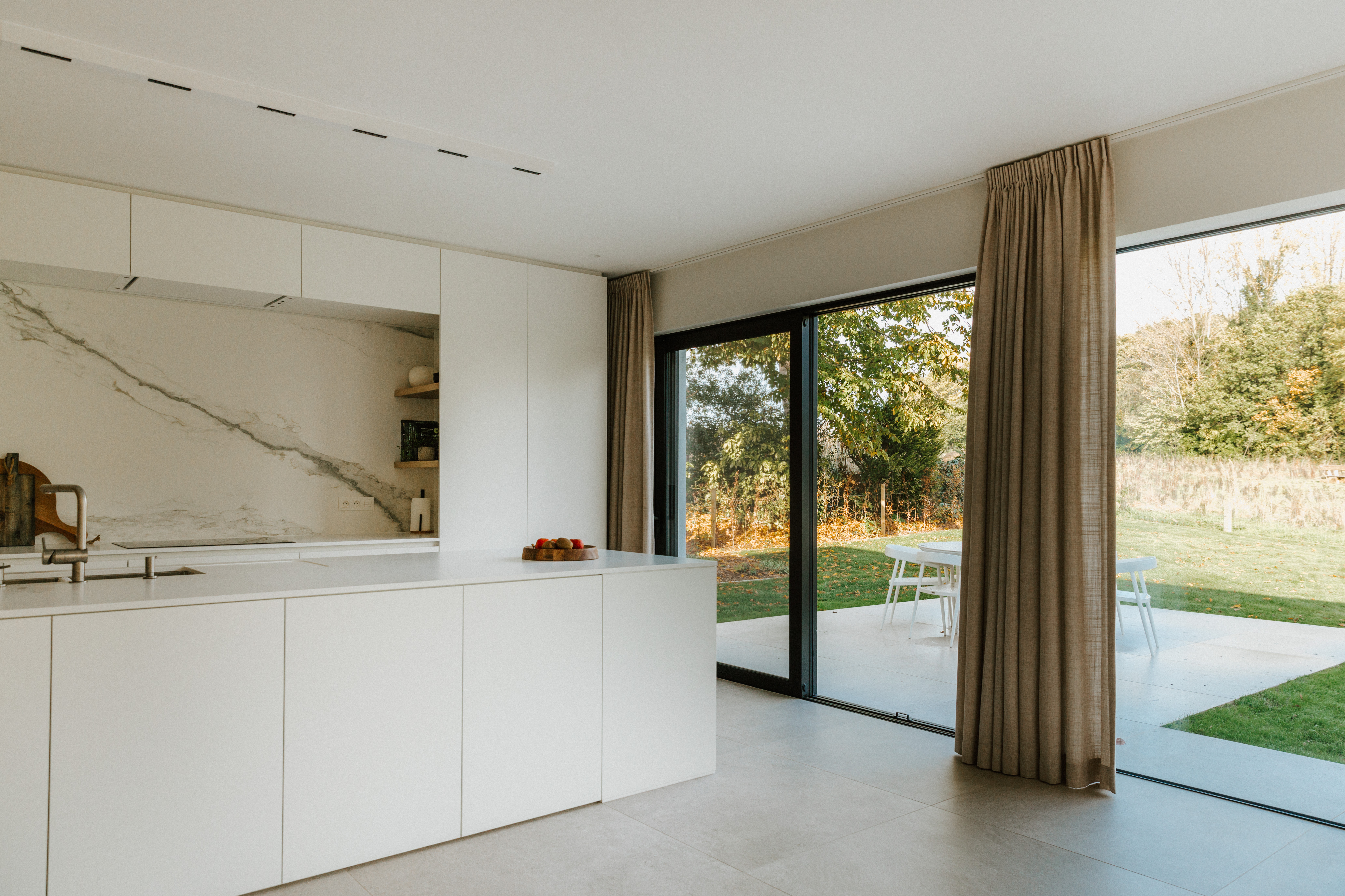 Moderne, gerenoveerde keuken met een groot vast raam in zwart aluminium, dat uitkijkt op het terras en de groene tuin.
