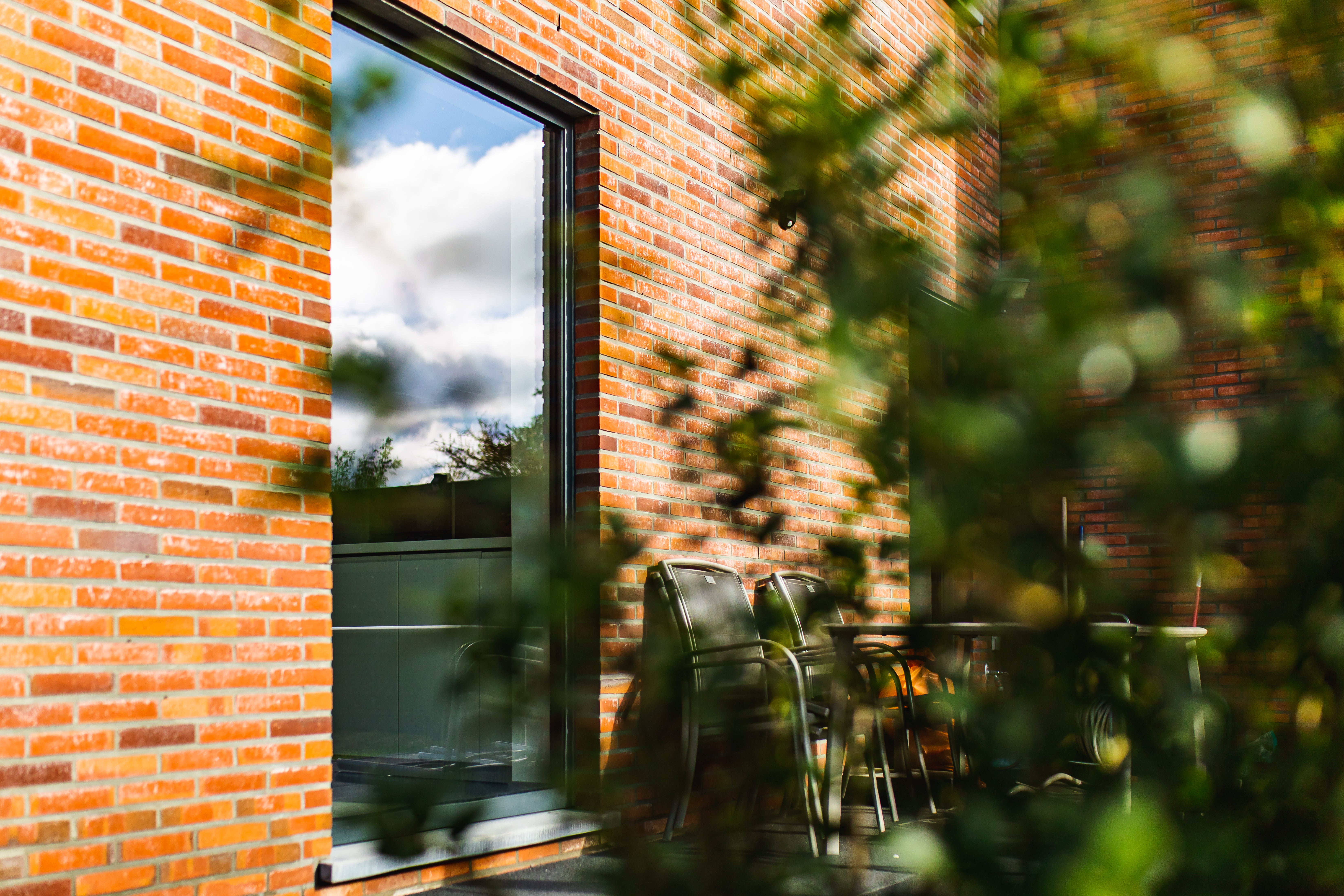 Hoog antracietgrijs raam in de bakstenen gevel van een vrijstaande nieuwbouwwoning, met tuinmeubelen op het terras en groen op de voorgrond.