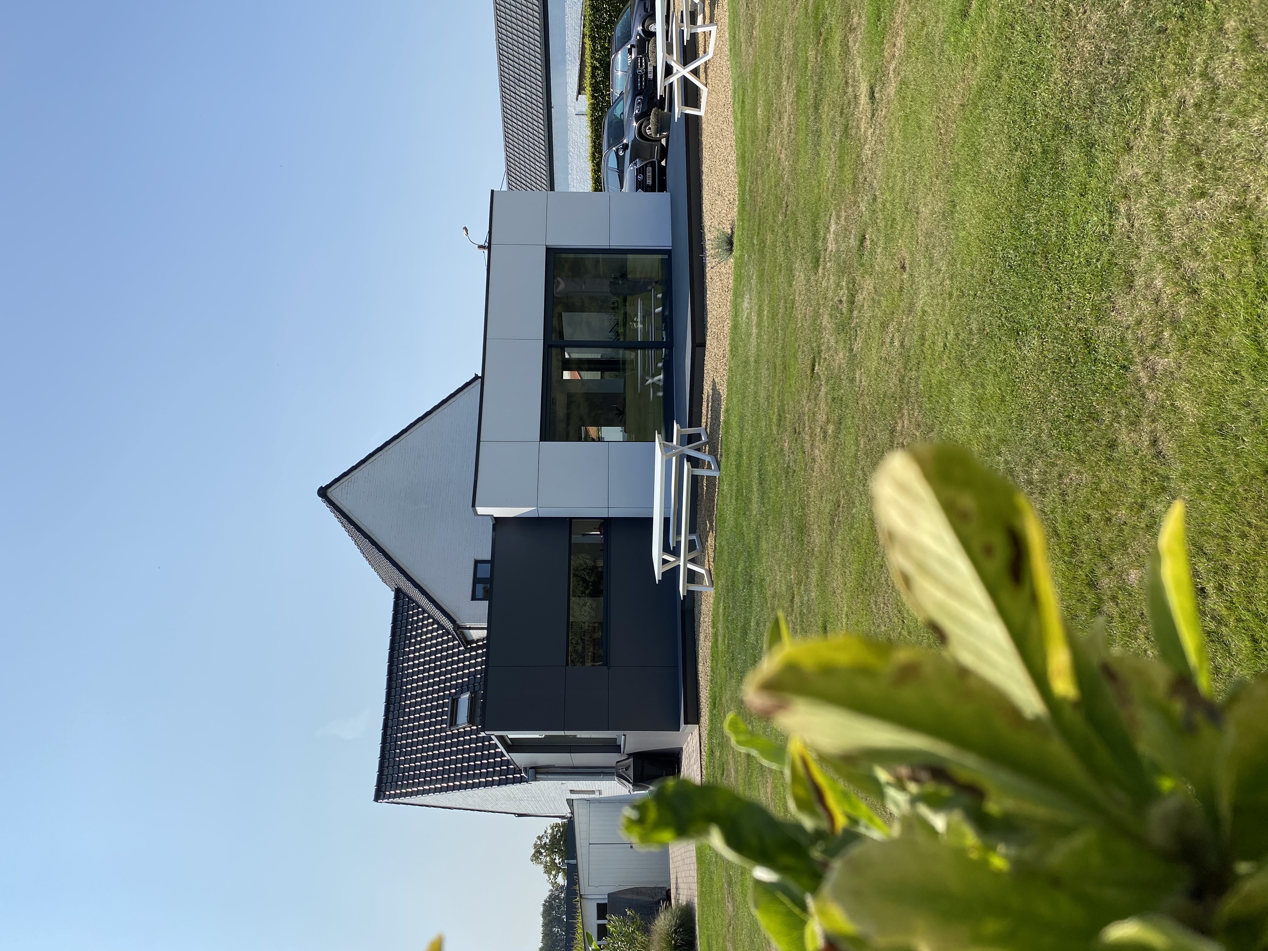 Moderne vrijstaande woning met donkere gevel en grote raampartijen, aan een grasveld met terras en picknicktafels.