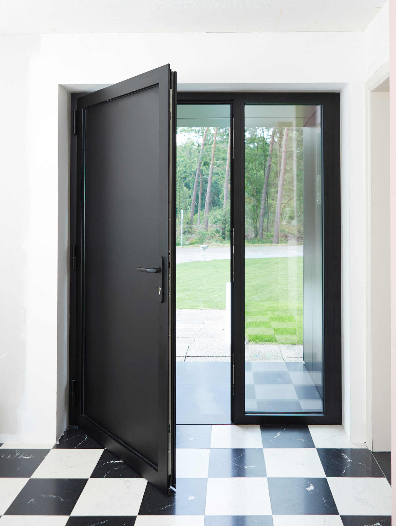 Zwarte steellookdeur met vast zijlicht in een karaktervolle, moderne inkomhal van een gerenoveerde open bebouwing, met zwart-witte tegels en zicht op de tuin.
