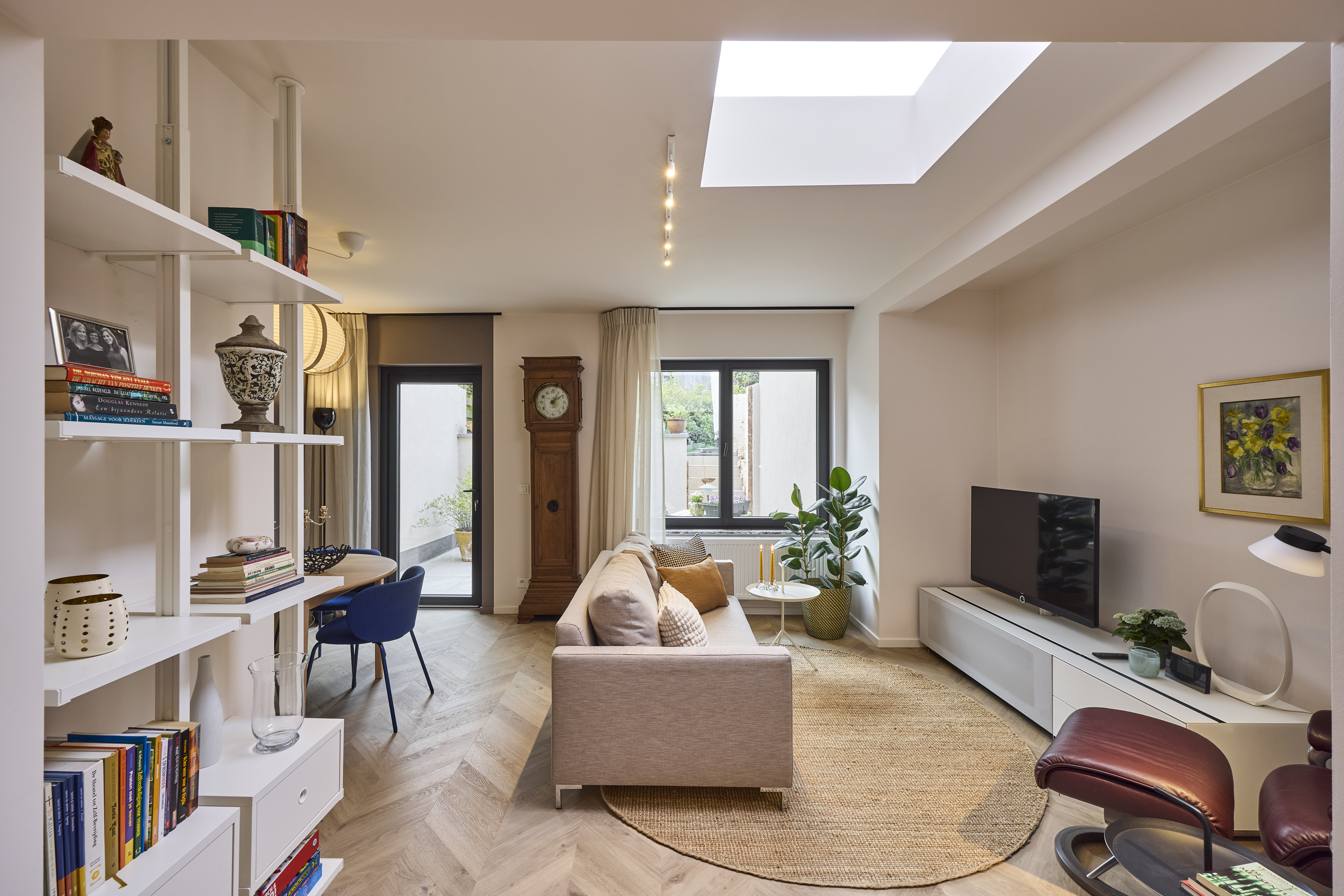 Lichte woonkamer met matzwart dubbel draai-kipraam, boekenkasten, sofa, televisie, vloerkleed en dakraam.