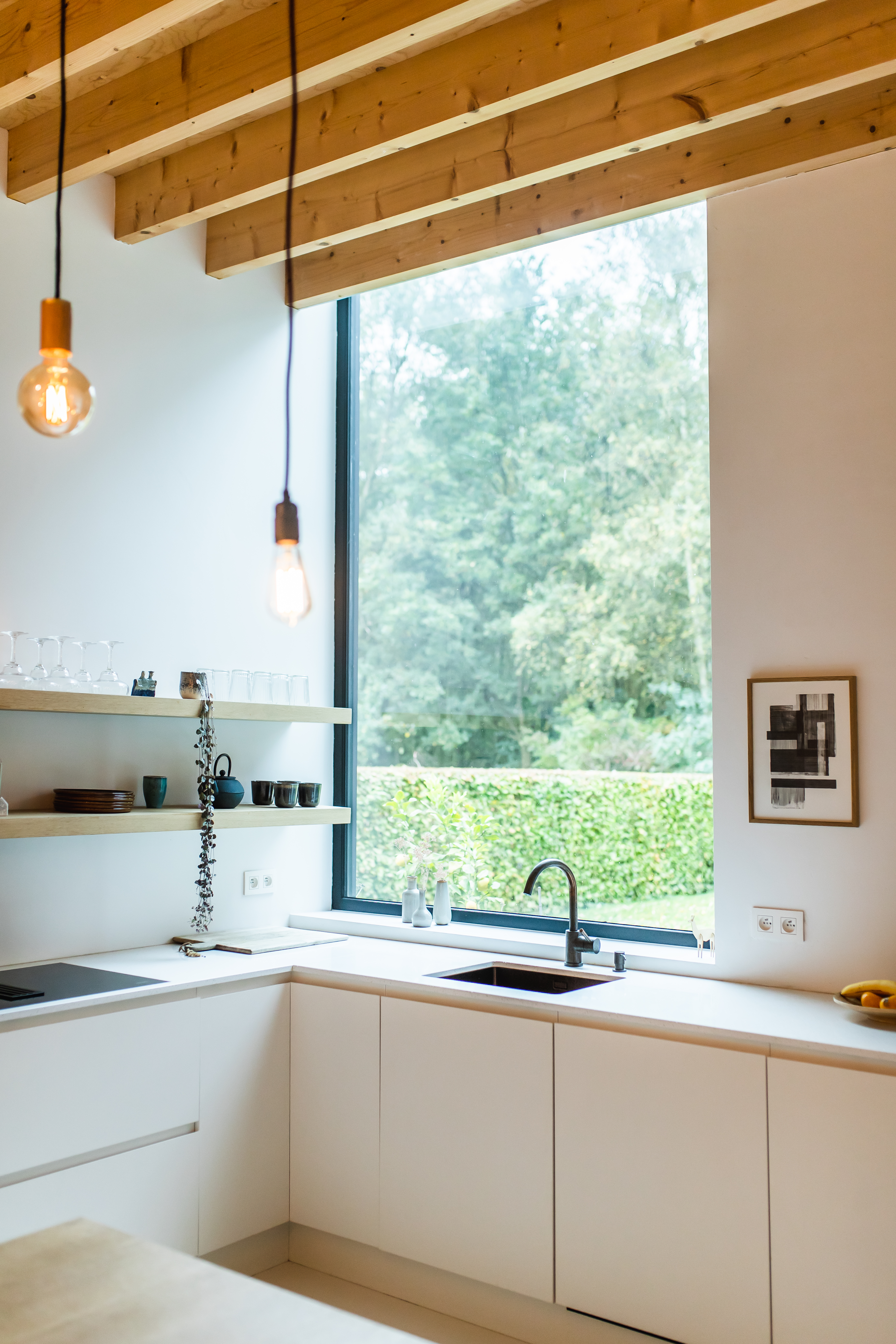 Moderne, witte keuken in een nieuwbouwwoning met een groot raam met zwarte 9005TC-profielen, een lichte houten draagstructuur en zicht op het groen.