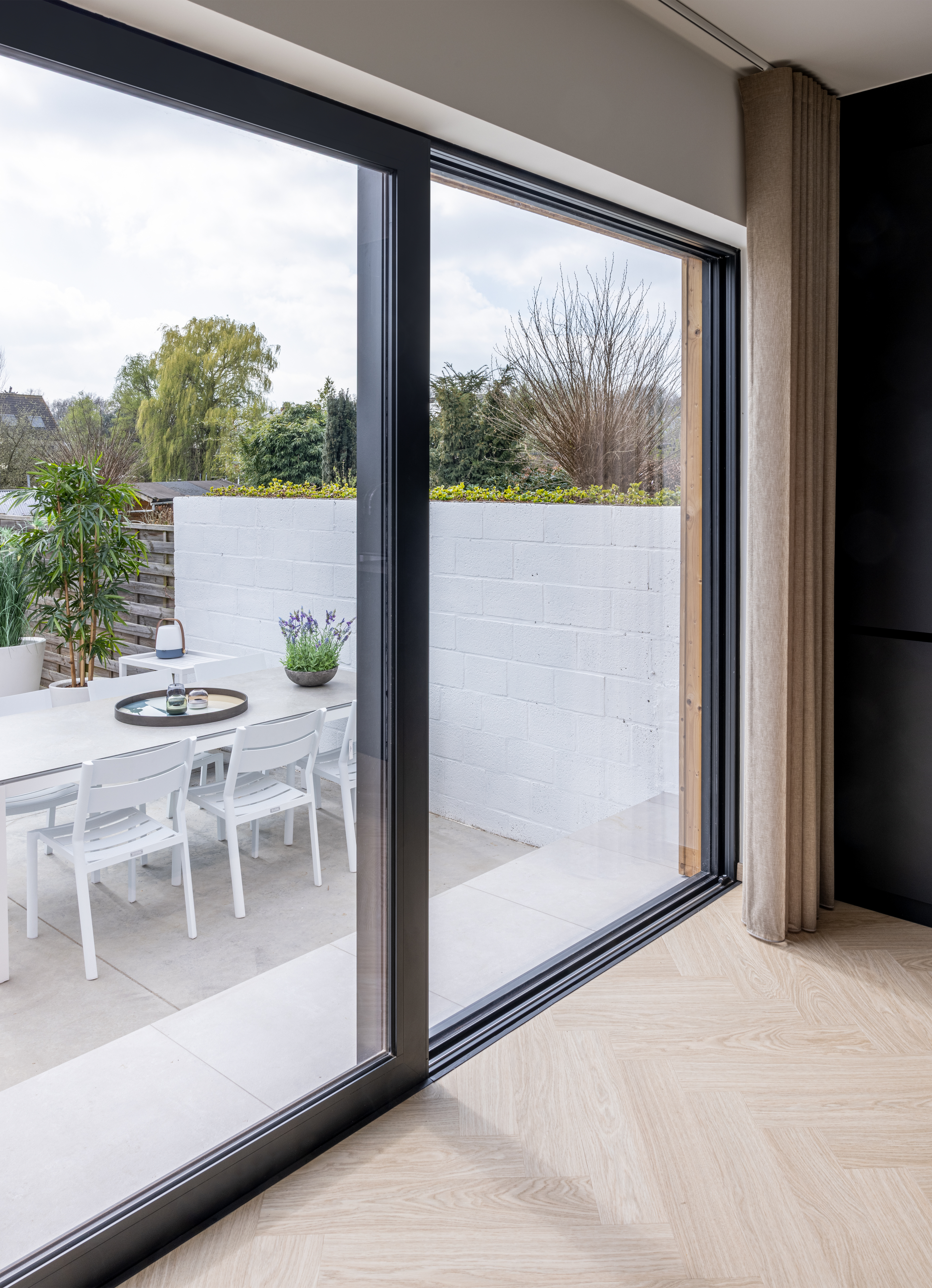 Groot donker schuifraam met zicht op een terras met witte tafel en stoelen, een lage muur en een tuin.