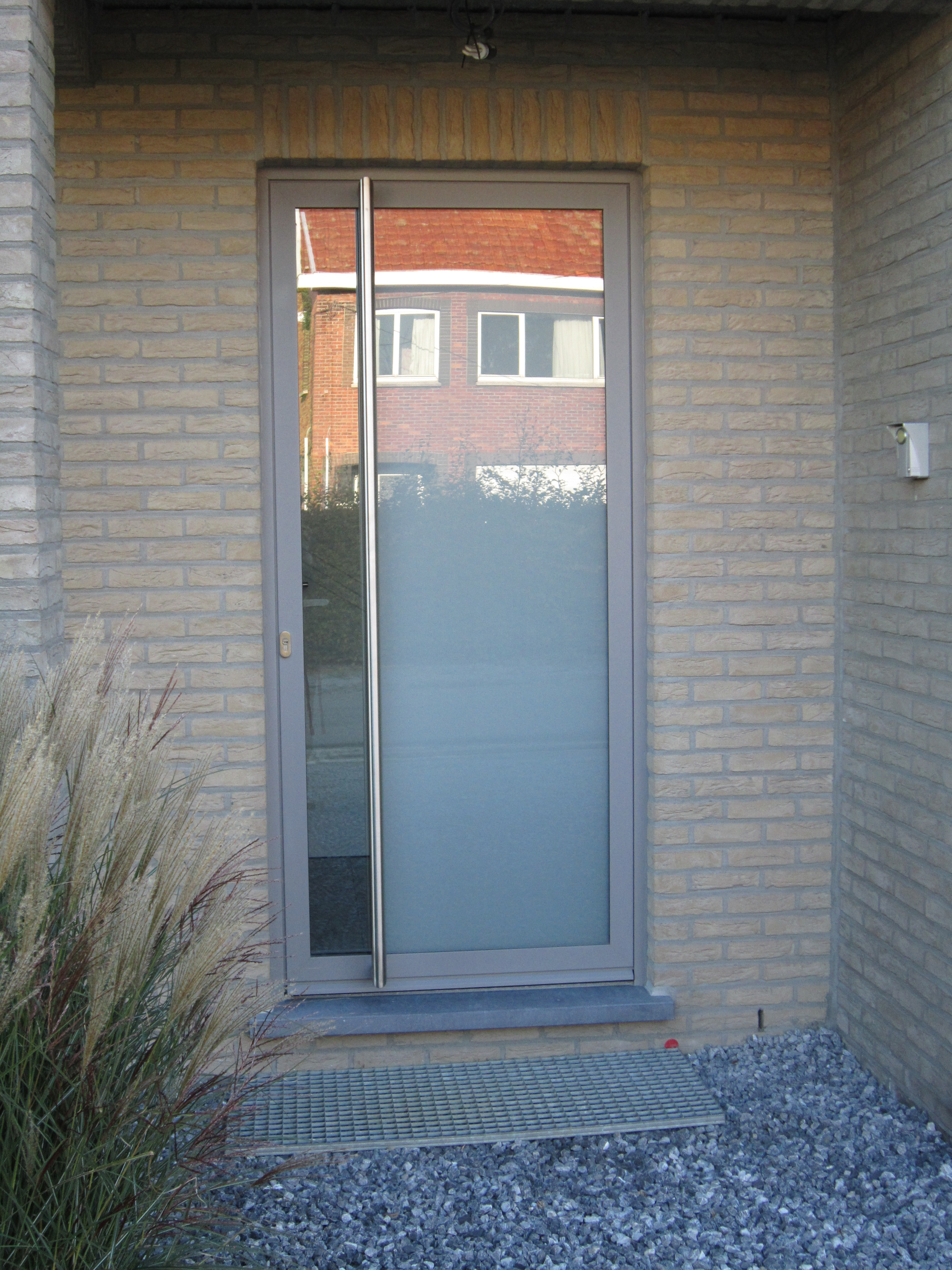 Porte d’entrée vitrée gris béton dans la façade en briques d’une maison quatre façades, avec un panneau latéral étroit et du gravier devant l’entrée.