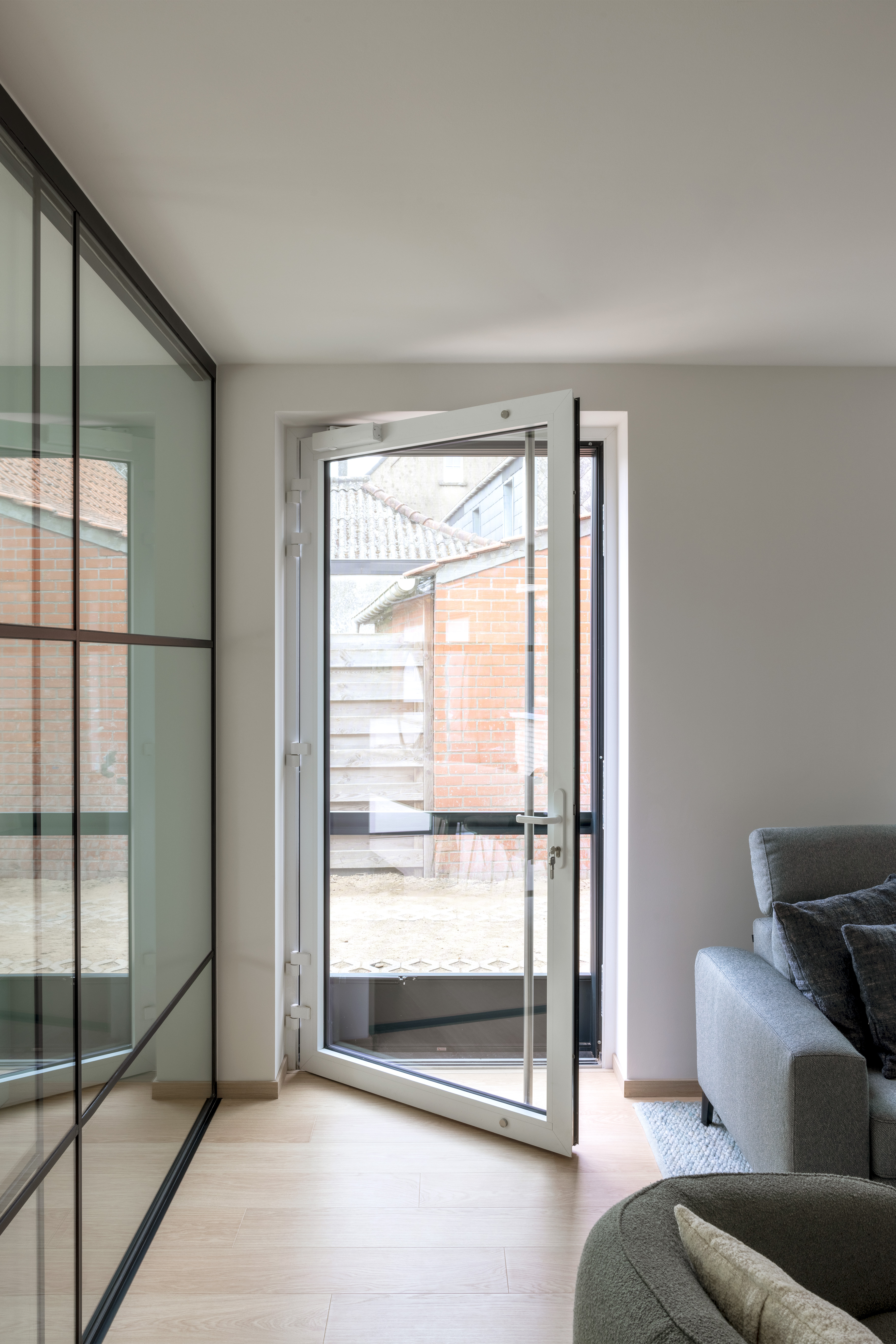 Een modern vormgegeven beglaasde vliegendeur met matwit aluminium raamprofiel staat open in een gerenoveerde woonkamer.