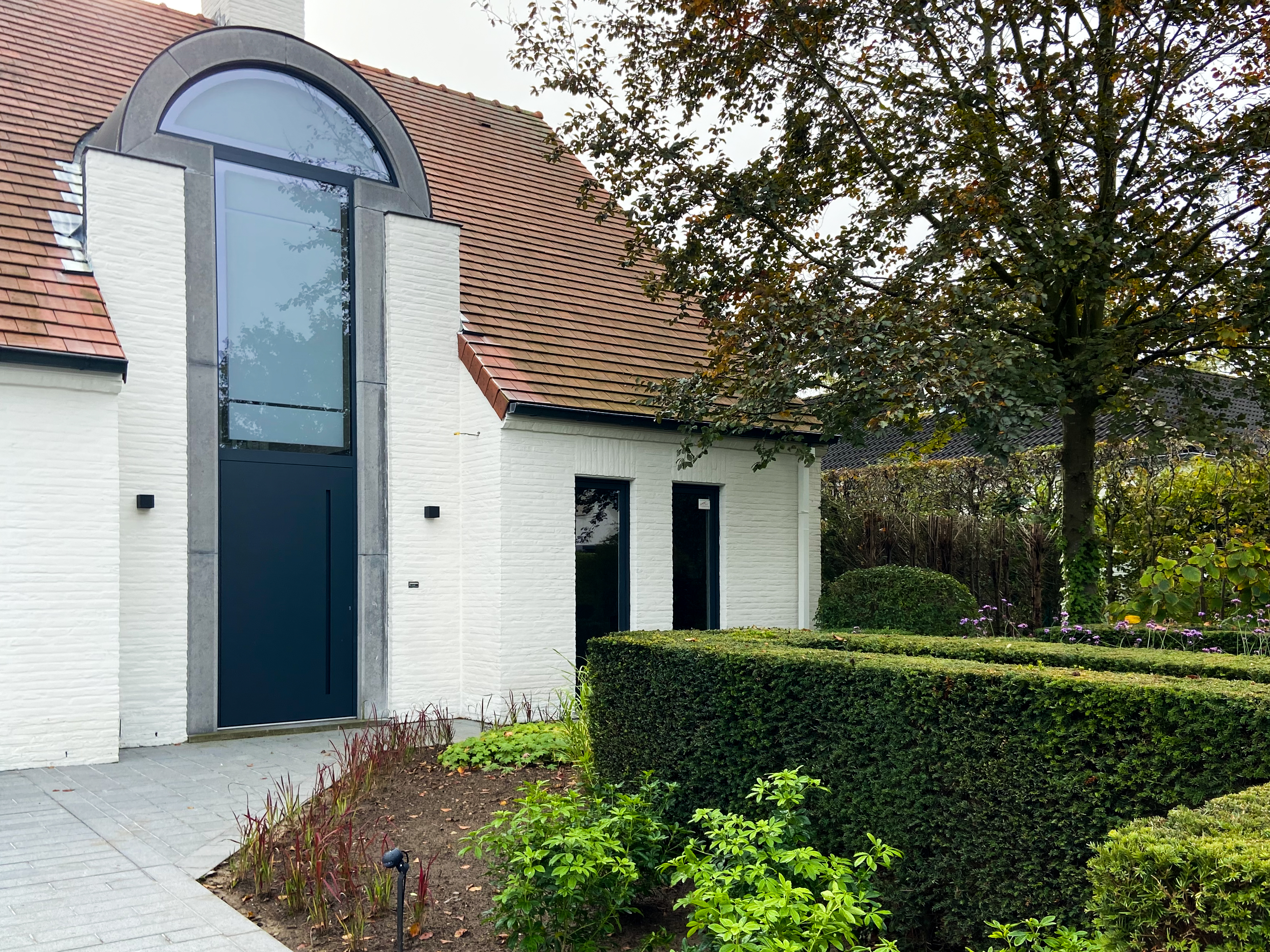 Gerenoveerde vrijstaande woning met witte gevel, hoge beglaasde voordeur met rondboog en verzorgde haag aan de voorzijde.