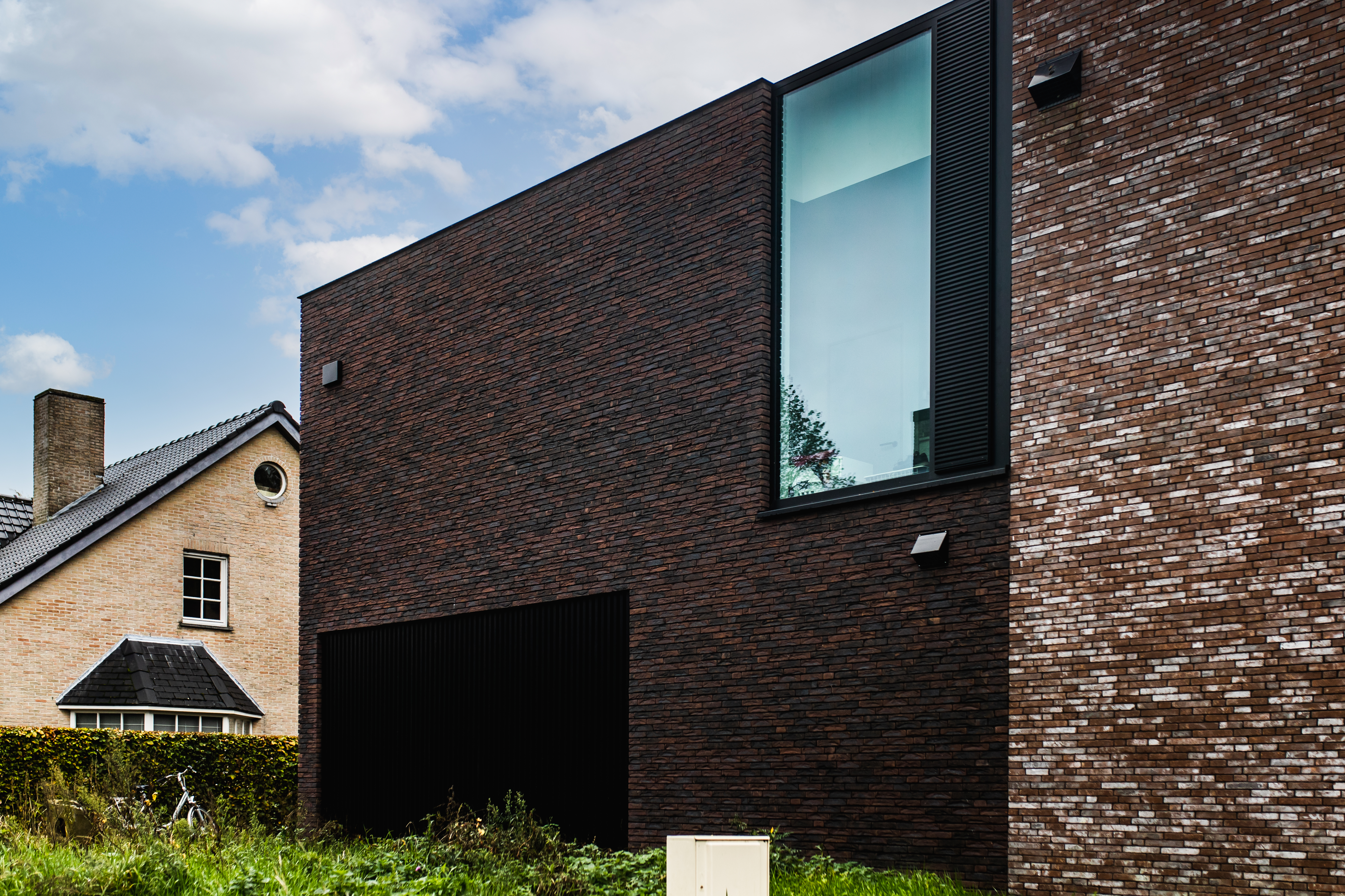 Karaktervolle moderne gevel van een nieuwbouwwoning in open bebouwing, met donkere baksteen, zwart 9005TC-raamprofiel, hoog raam en zwarte garagepoort.