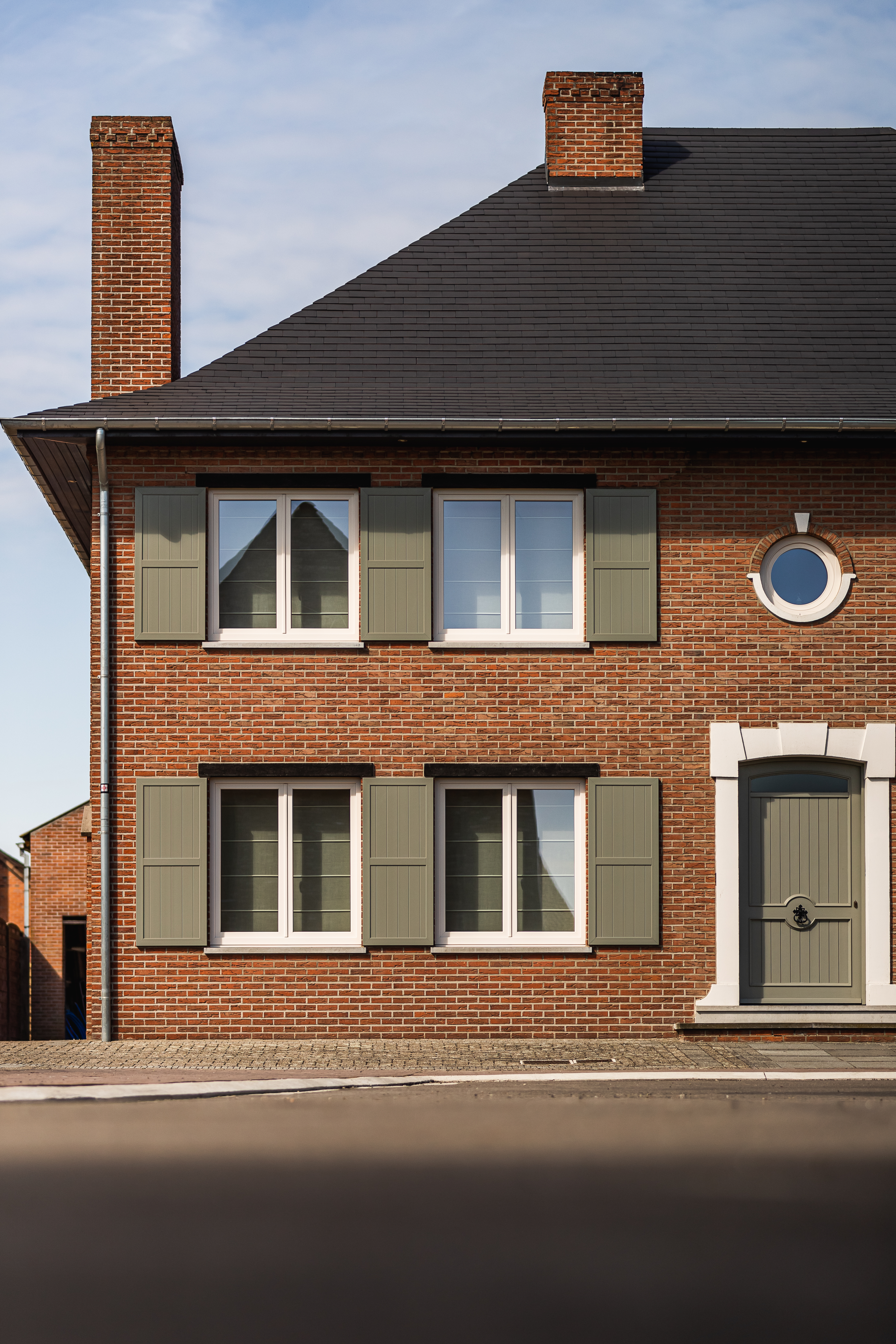 Gerenoveerde vrijstaande bakstenen woning met crèmekleurige dubbele ramen, groene klapluiken, een donker dak, schoorstenen en een grijze voordeur.