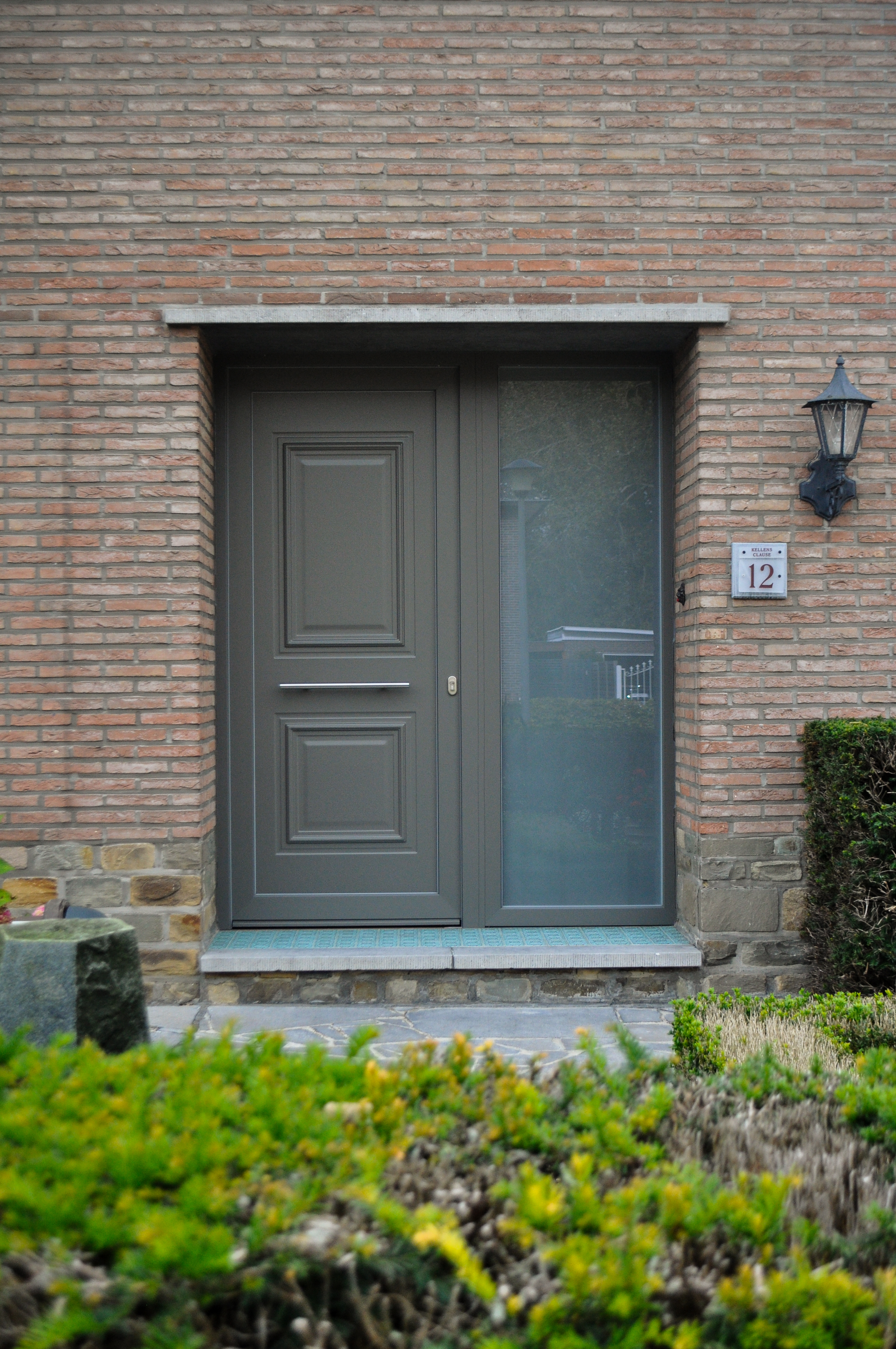 Kwartsgrijze PVC-voordeur met panelen en een smal zijlicht in de gerenoveerde bakstenen gevel van een halfopen woning.