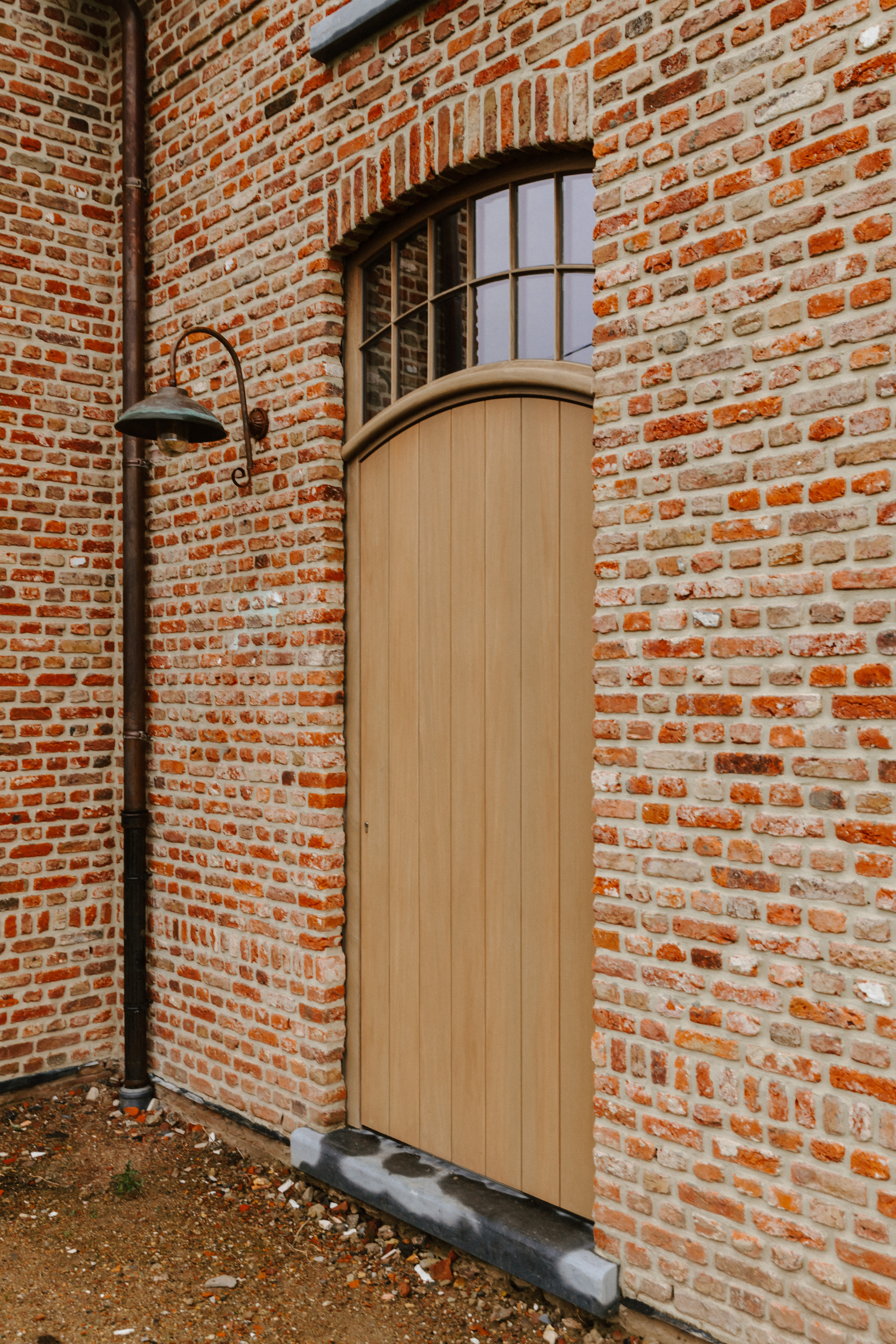 Hoge deur in natuurlijk Afrormosia met bovenlicht, geïntegreerd in een warme, landelijke bakstenen gevel van een vrijstaande nieuwbouwwoning.