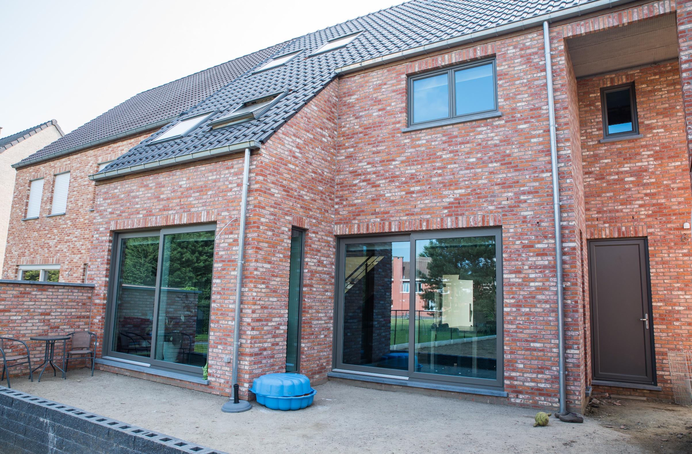Tijdloze halfopen nieuwbouwwoning met rode bakstenen gevel, grote ramen en kwartsgrijze NERF-raamprofielen aan de voorzijde.